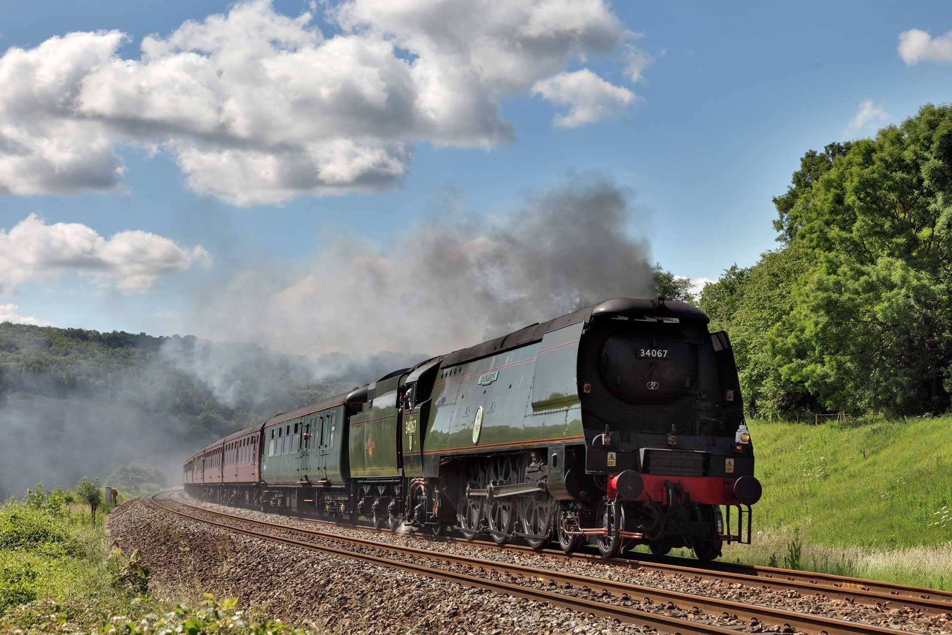 Steam train, black and green, traveling on tracks through a green landscape under a cloudy blue sky.