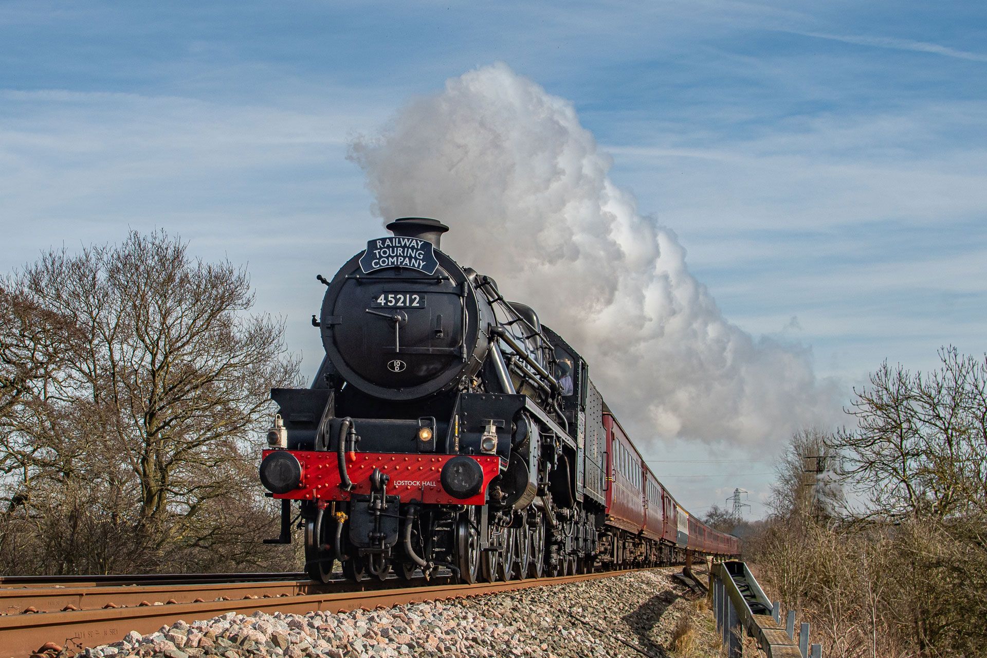 Black steam locomotive pulling cars through a rural landscape, with white smoke billowing behind it.