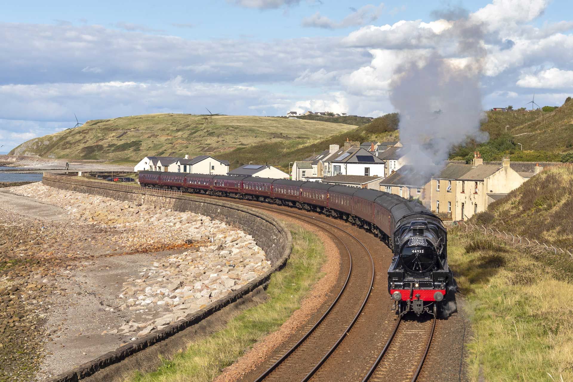 Steam train rounding a bend by the coast, puffing white smoke. Brown tracks, stone wall, houses, and hillside in view.