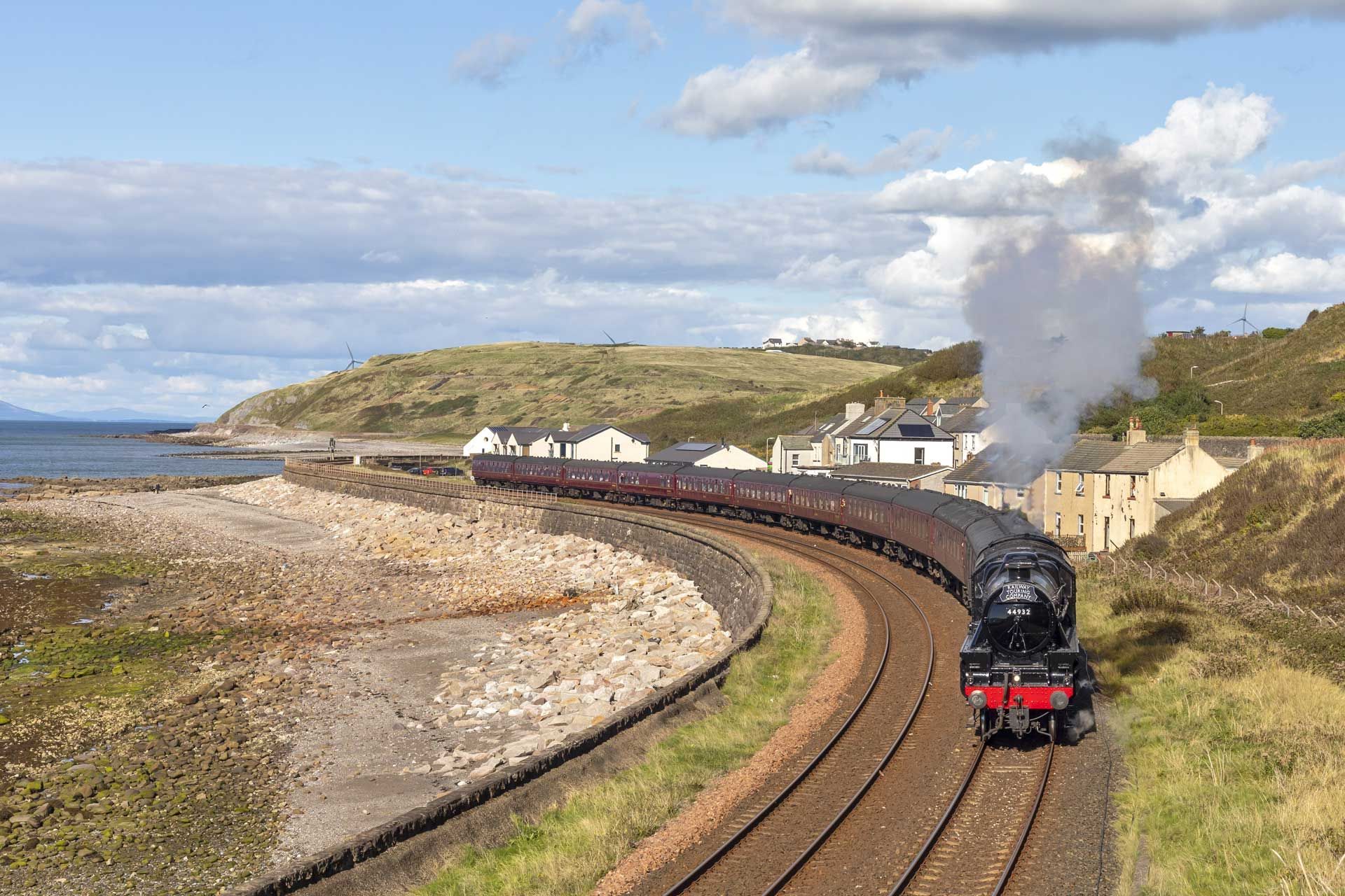 Steam train curving along coastal tracks, passing white buildings under a blue sky.