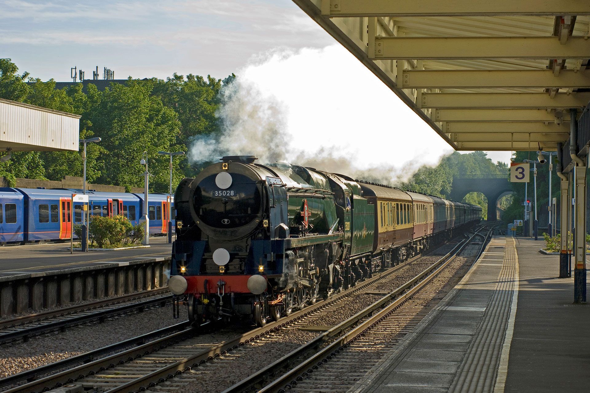 Steam locomotive at a train station platform, billowing white smoke beside waiting passenger cars.