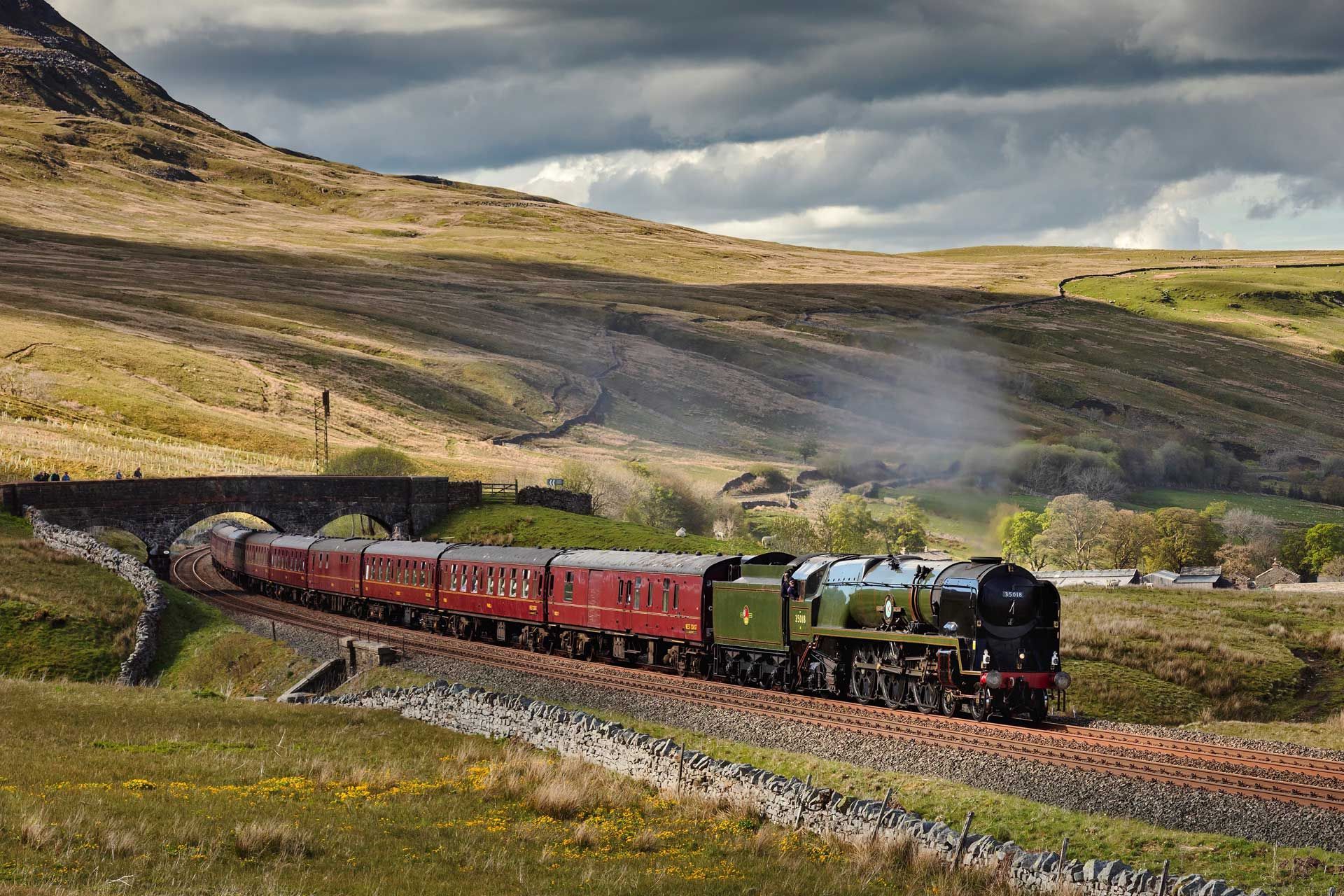 Steam train traveling through a rural landscape, emerging from a tunnel. Green and red carriages.