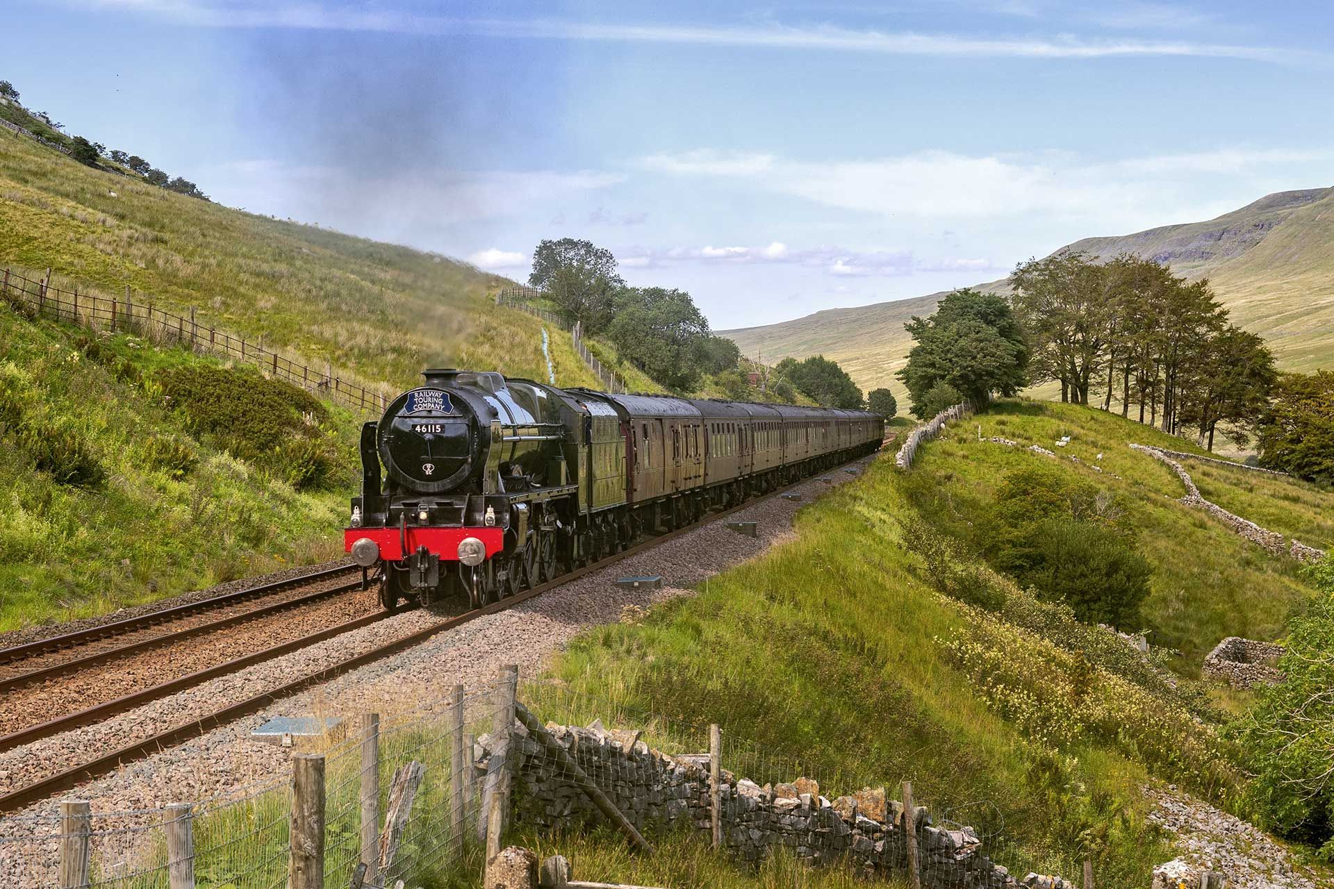 Steam train traveling through a green valley with a stone wall and blue sky.