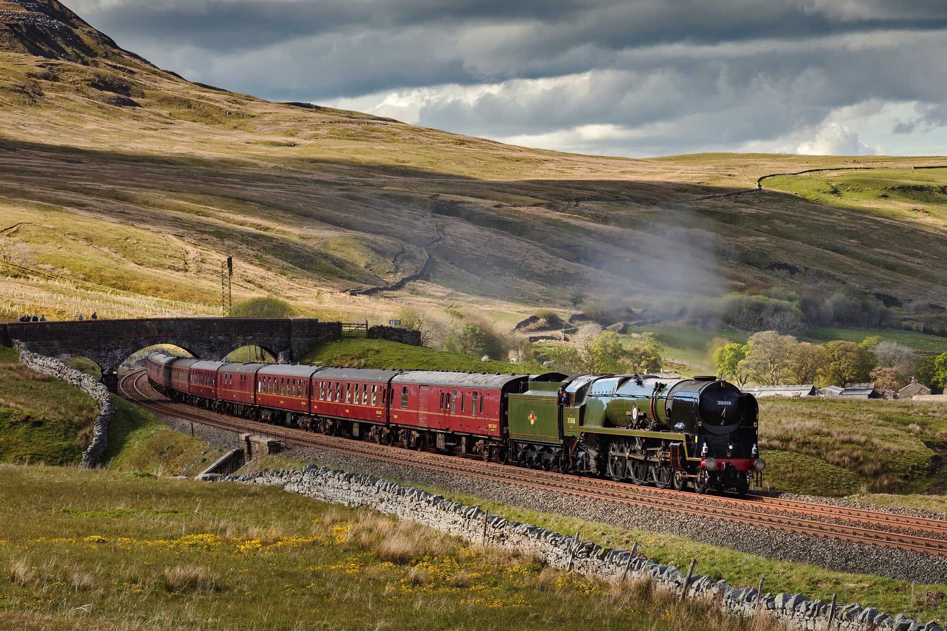 Steam train traveling along tracks through a hilly, grassy landscape, passing under a stone bridge.