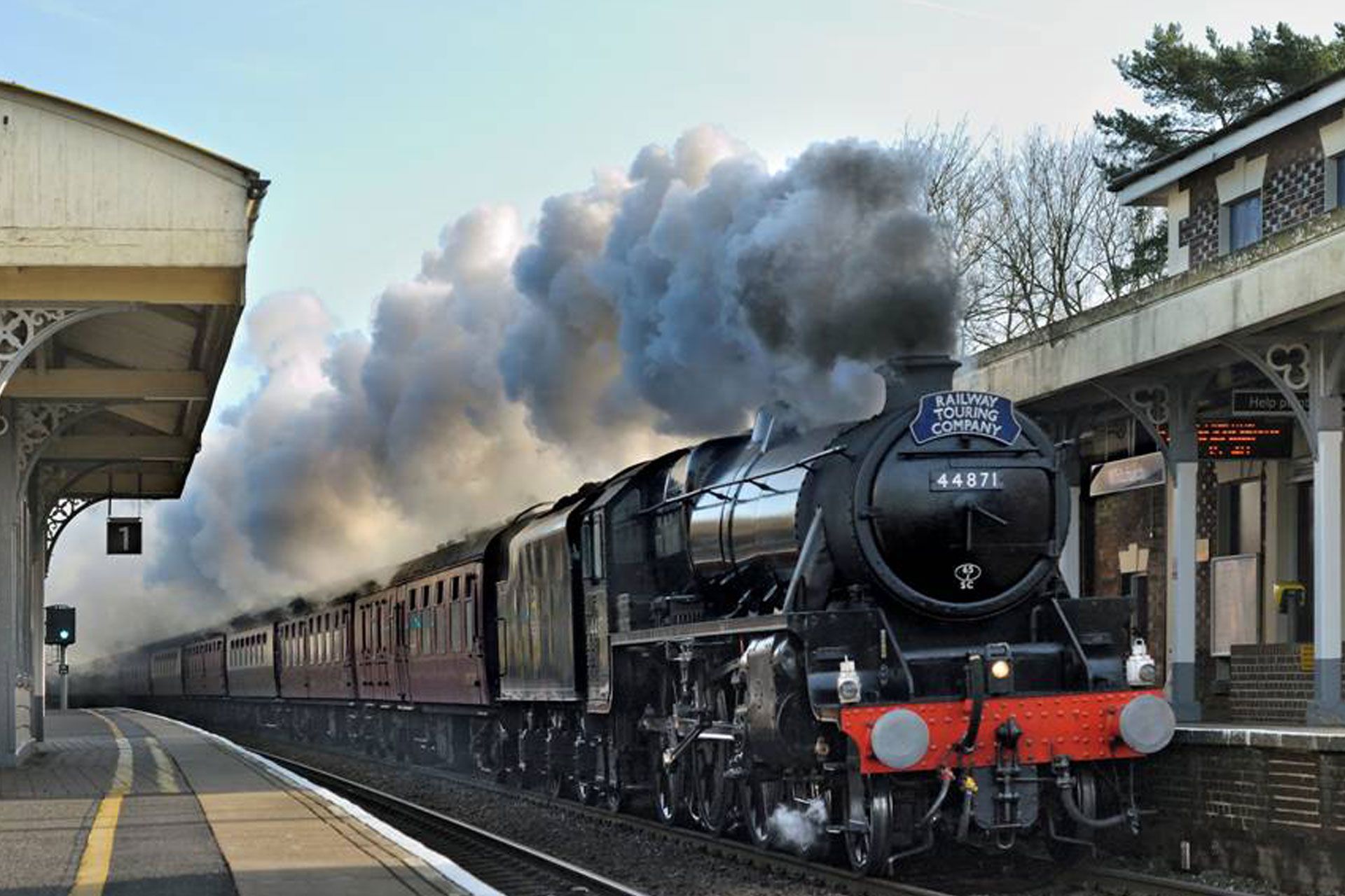 Black steam locomotive pulling a train through a station, billowing smoke under a cloudy sky.
