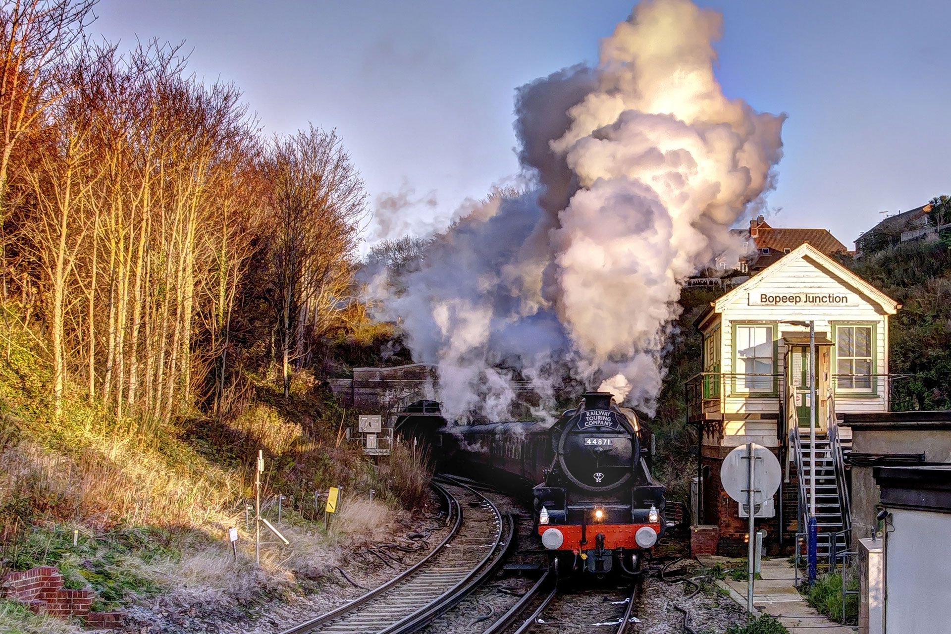Steam train on curving tracks beside a small station, with a tall plume of white smoke at sunset.