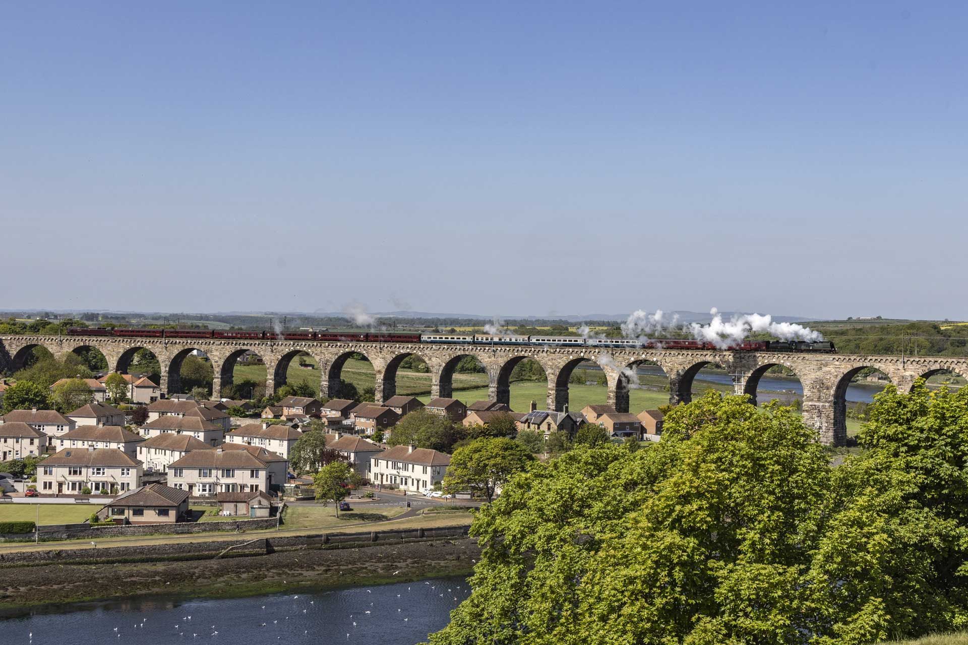 Steam train crossing a large stone viaduct over a town and river on a sunny day.