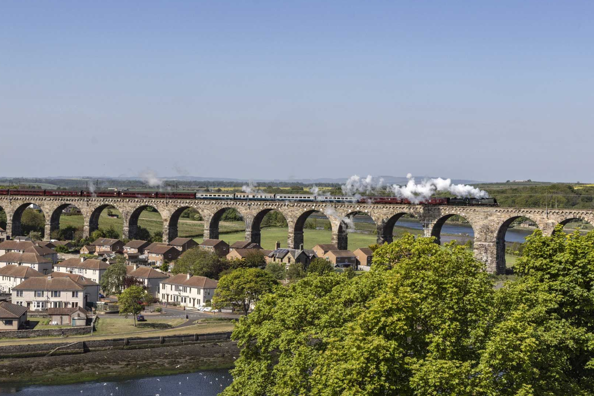 A steam train crossing a stone railway viaduct above a small town and river on a clear, sunny day.