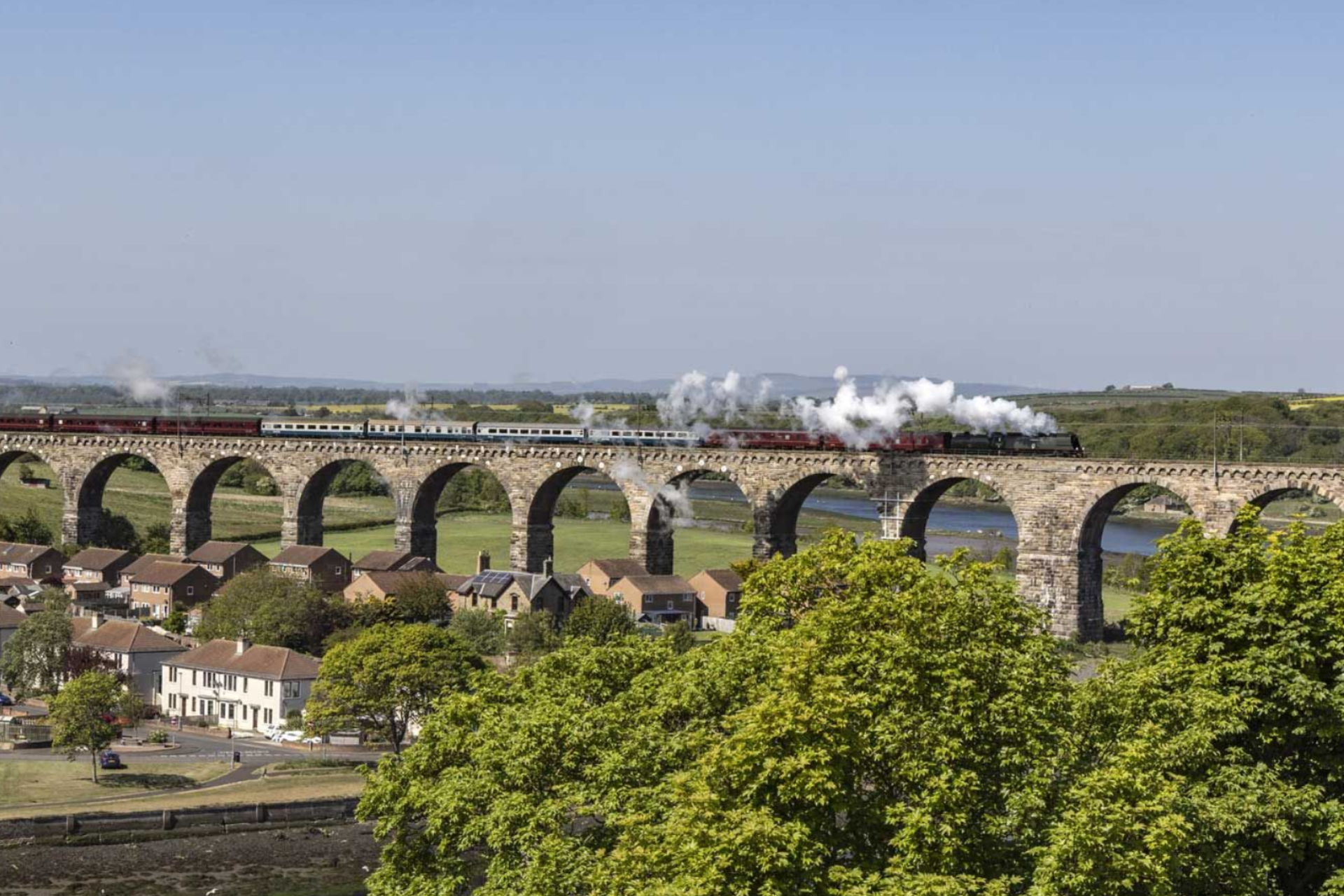 Steam train crossing a stone viaduct over a river, smoke billowing. Houses and trees in foreground.