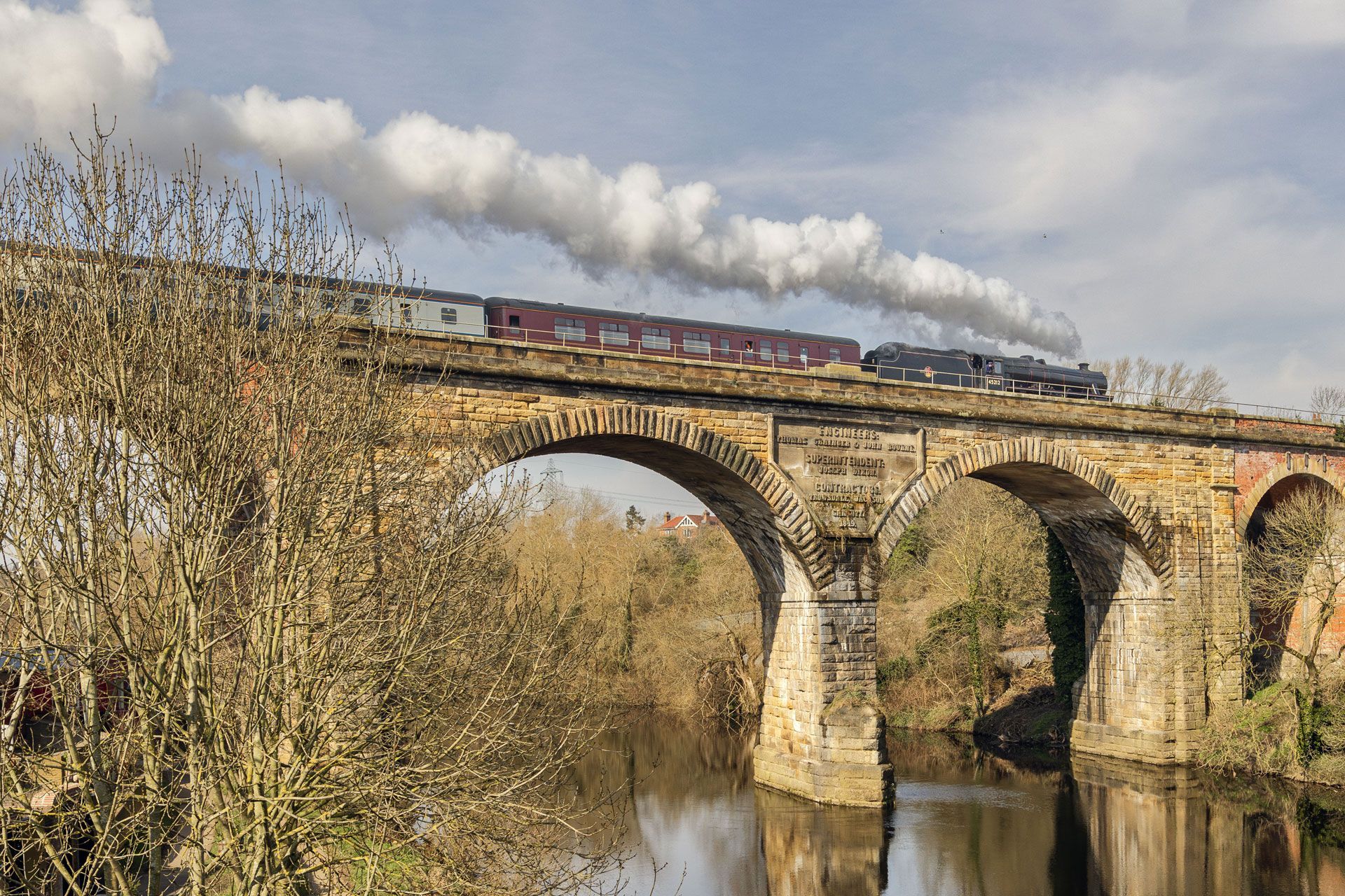 Steam train crossing a stone viaduct over a river, with trees and cloudy sky.