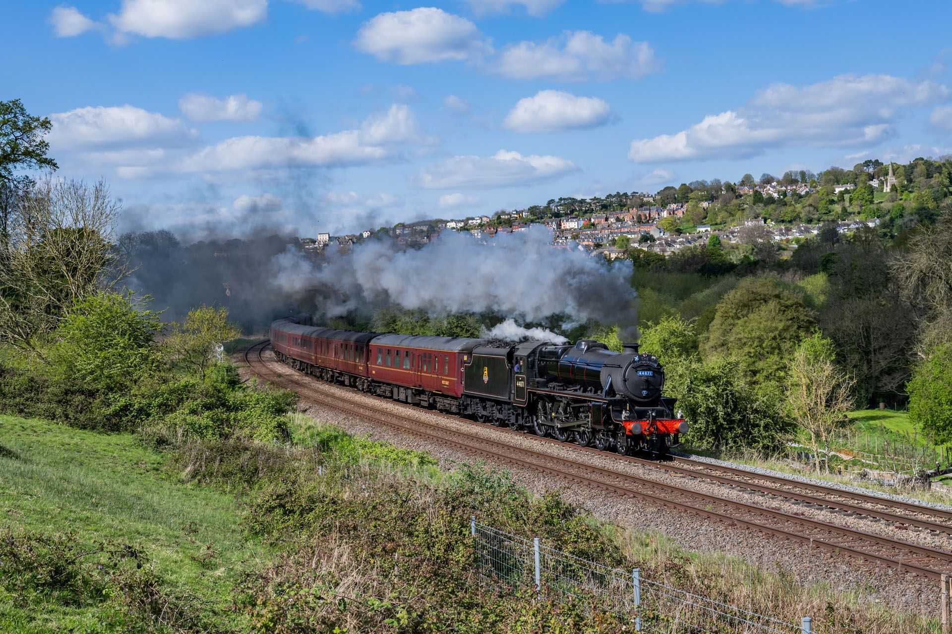 Steam train traveling on tracks through a green landscape, smoke billowing. Houses on a hillside in the background.