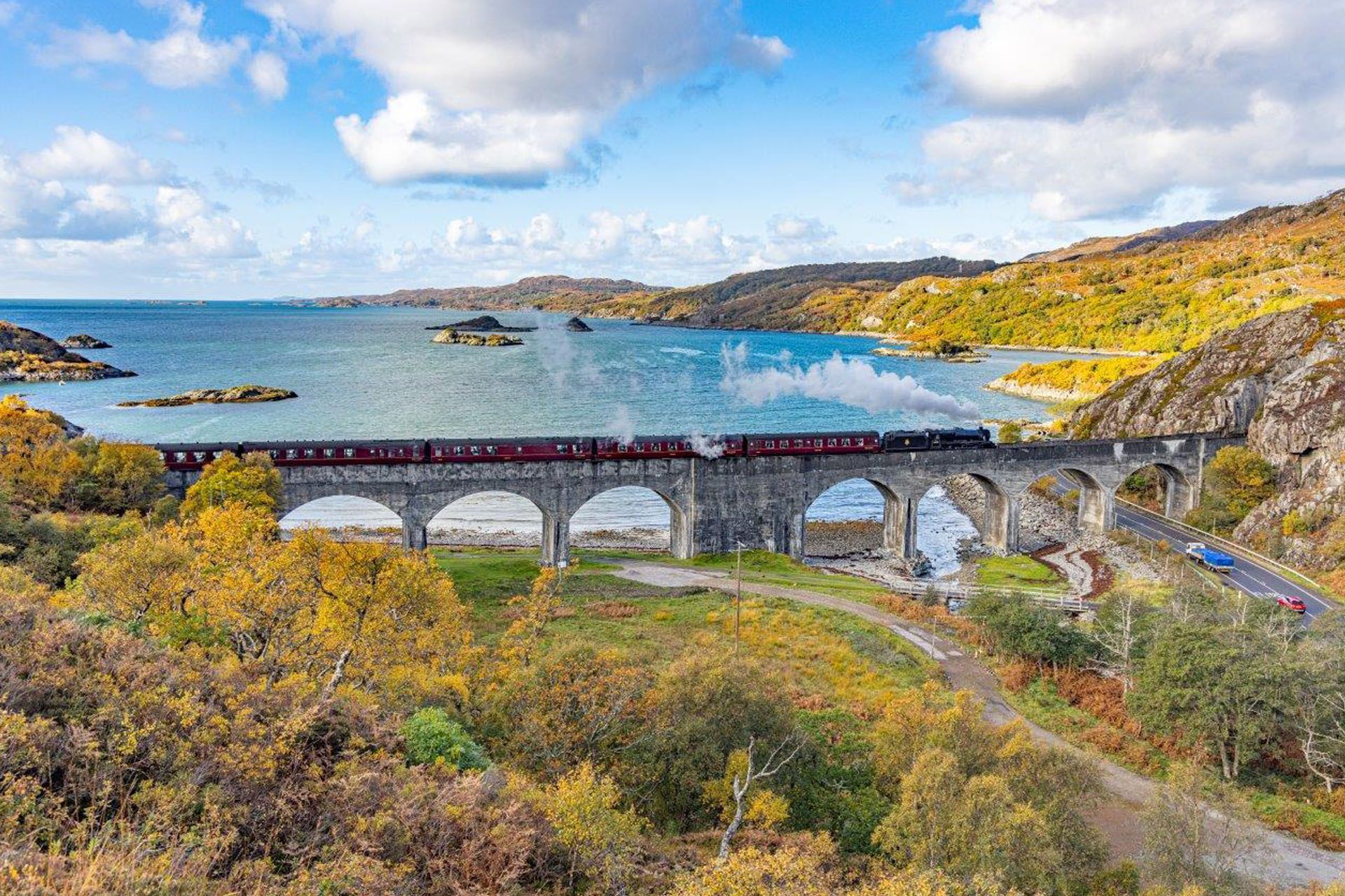 Train crossing the stone arches of a viaduct over water, with a forested hillside and blue sky in the background.