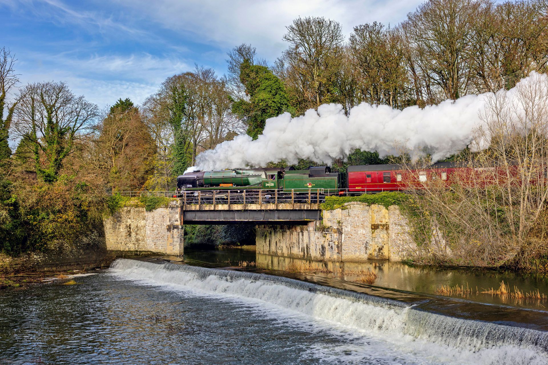 Steam train crossing a stone bridge over a river, with white smoke billowing.