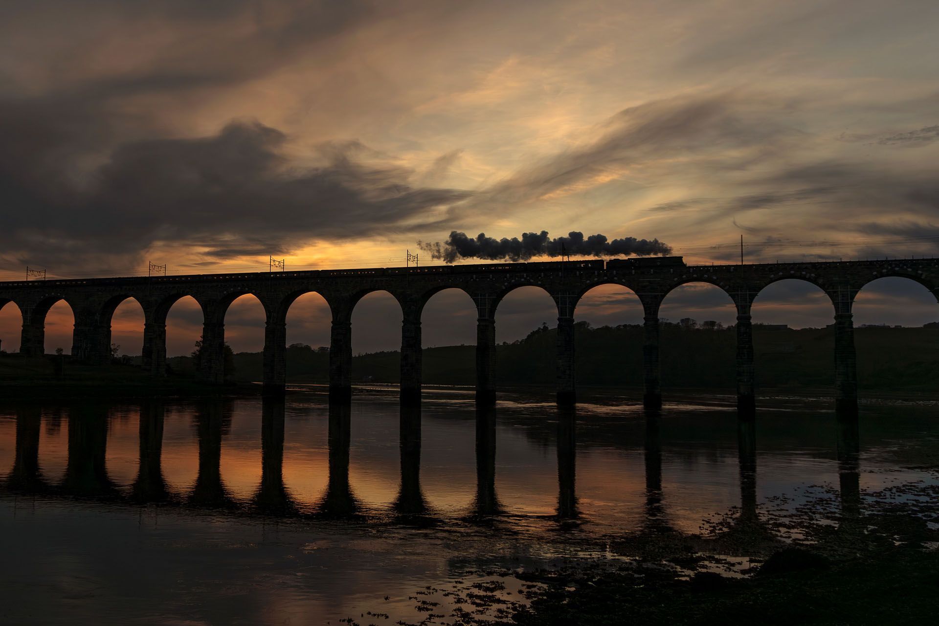 Train crossing a stone viaduct over a river at sunset; orange and gray sky reflects in the water.