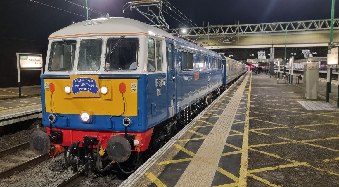 Diesel train on tracks, brown and yellow, surrounded by green trees and foliage, under a cloudy sky.