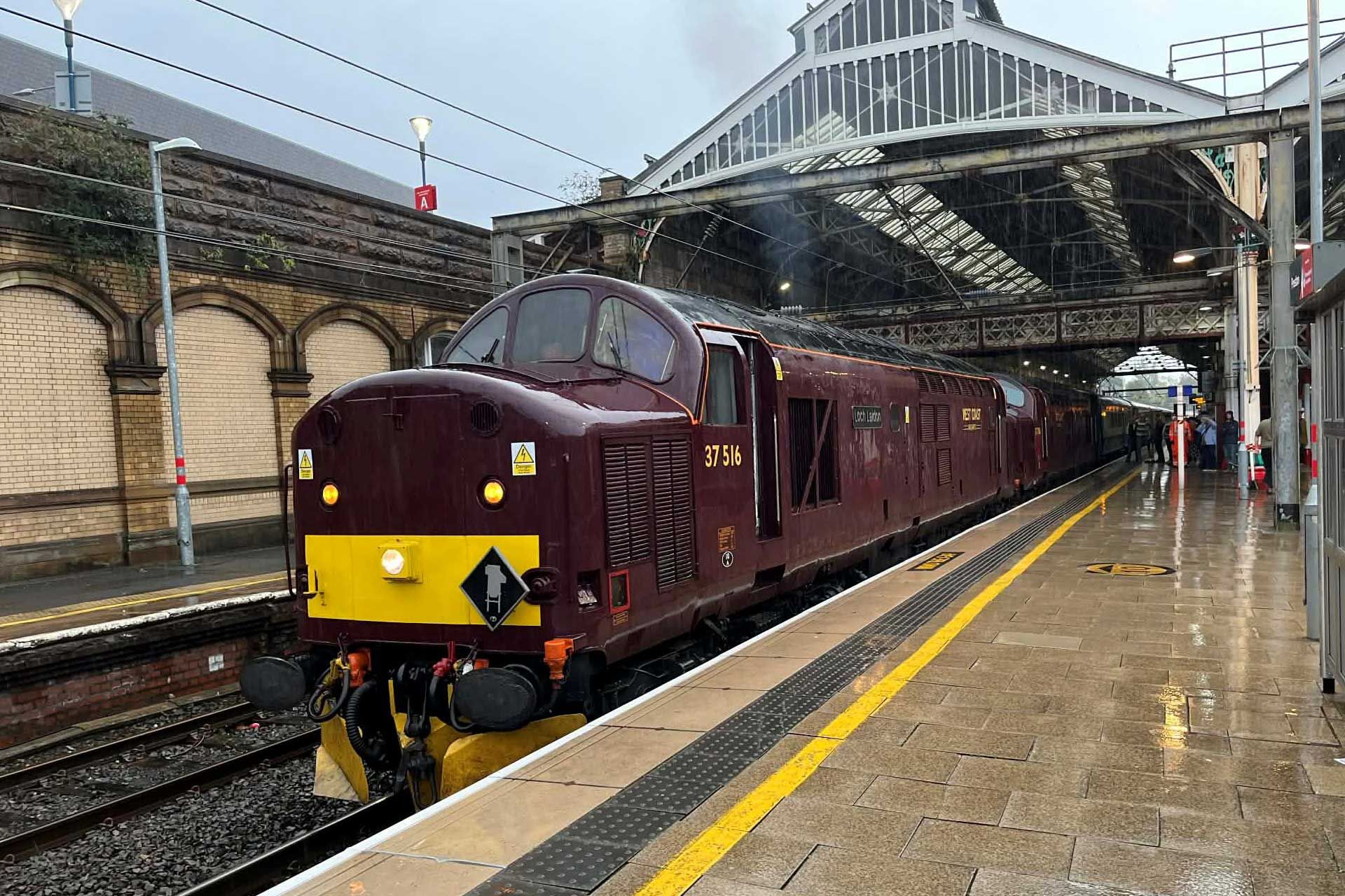 Maroon train at a station platform. Wet pavement and a glass-roofed station structure.