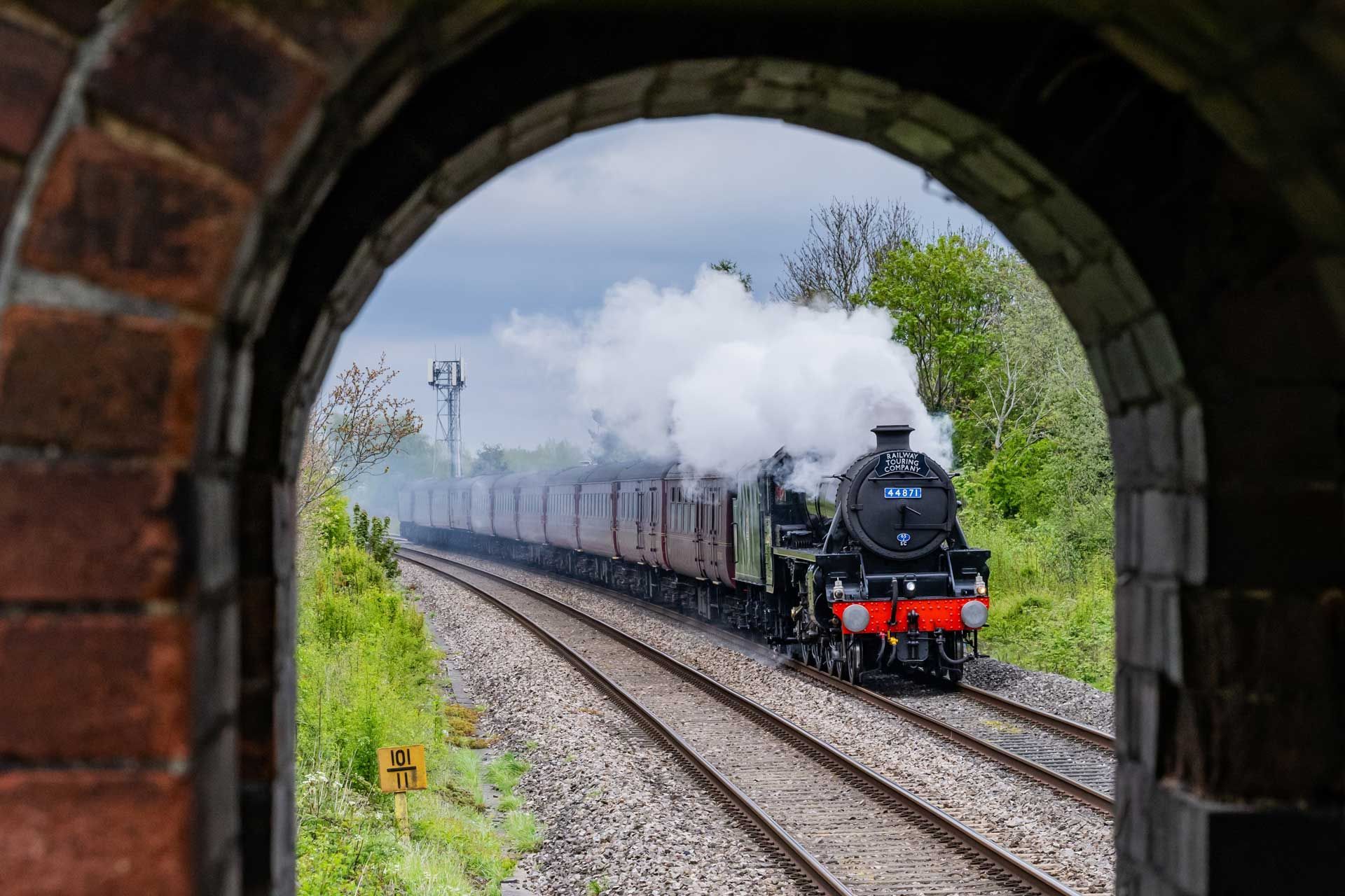 Steam train emerging from brick archway on tracks; plume of smoke.