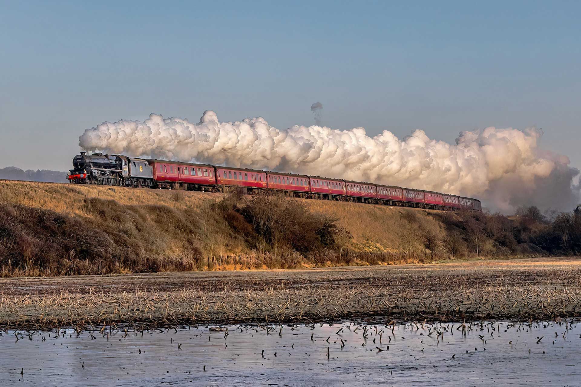 Steam train traveling on a track, emitting large plumes of white smoke against a clear blue sky.