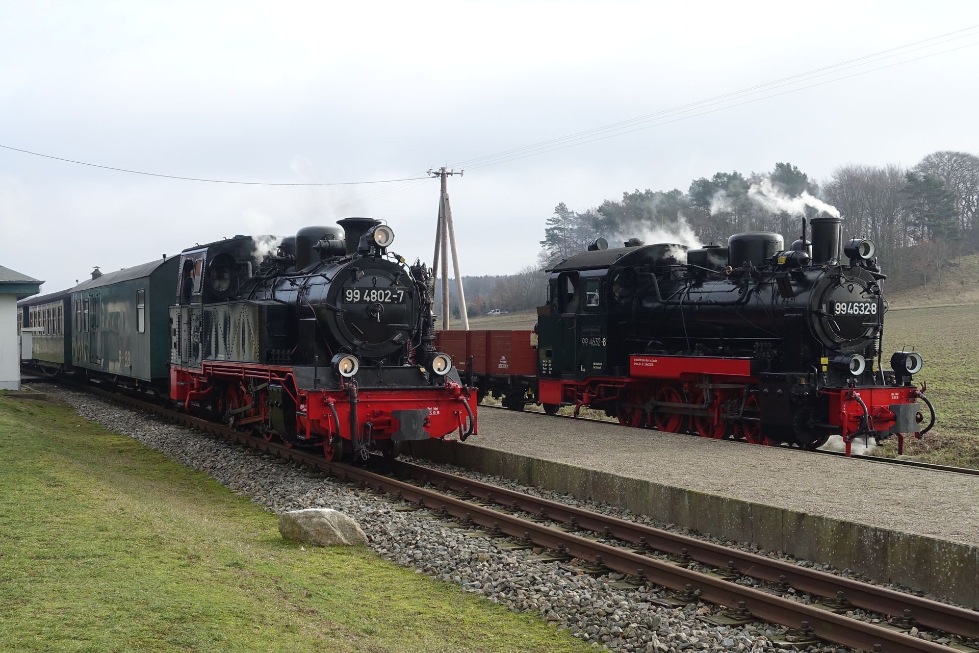 Two black steam locomotives with red wheels parked on tracks beside a platform under a cloudy sky