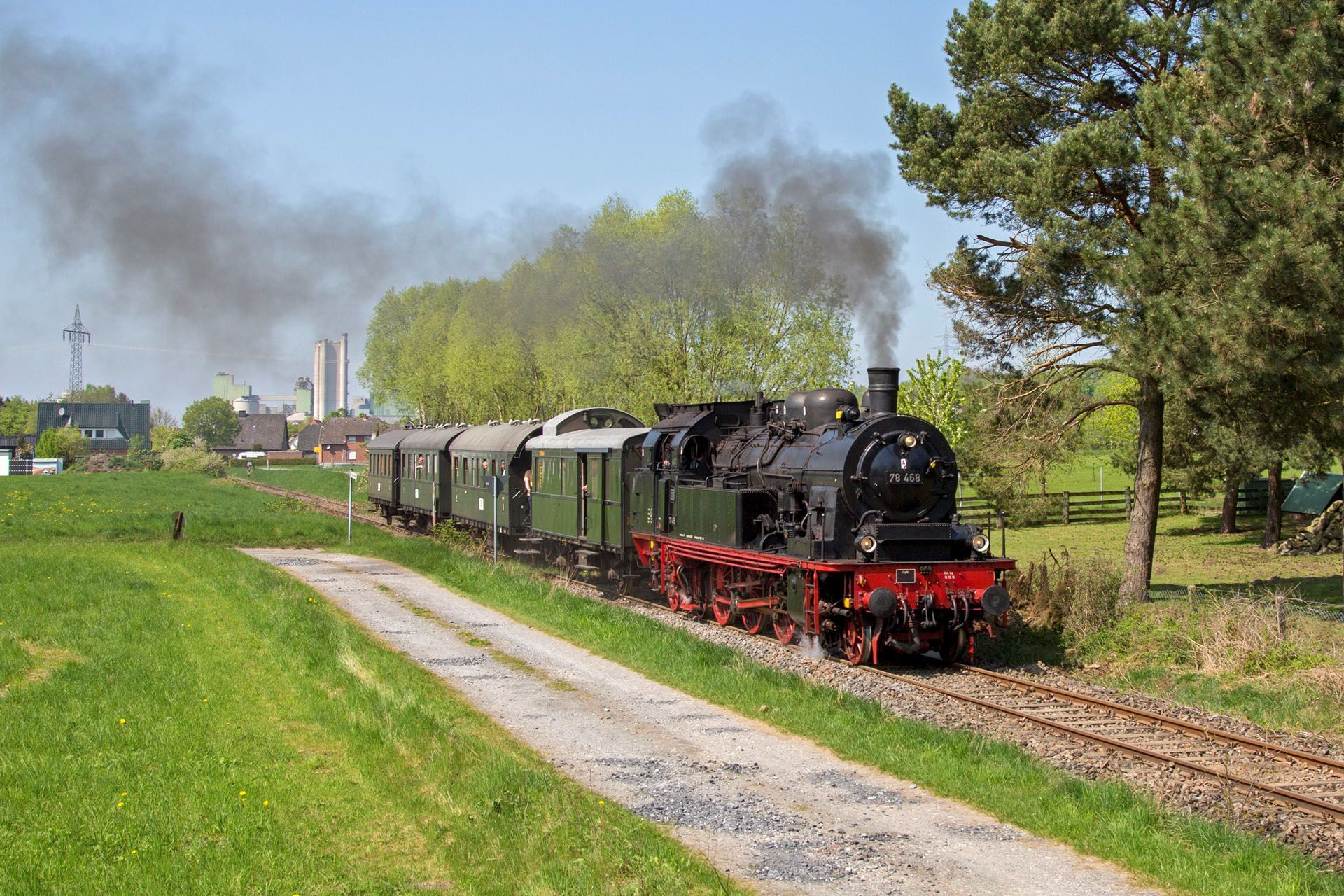 Steam train with black locomotive and green cars traveling through a sunny rural park, smoke drifting 