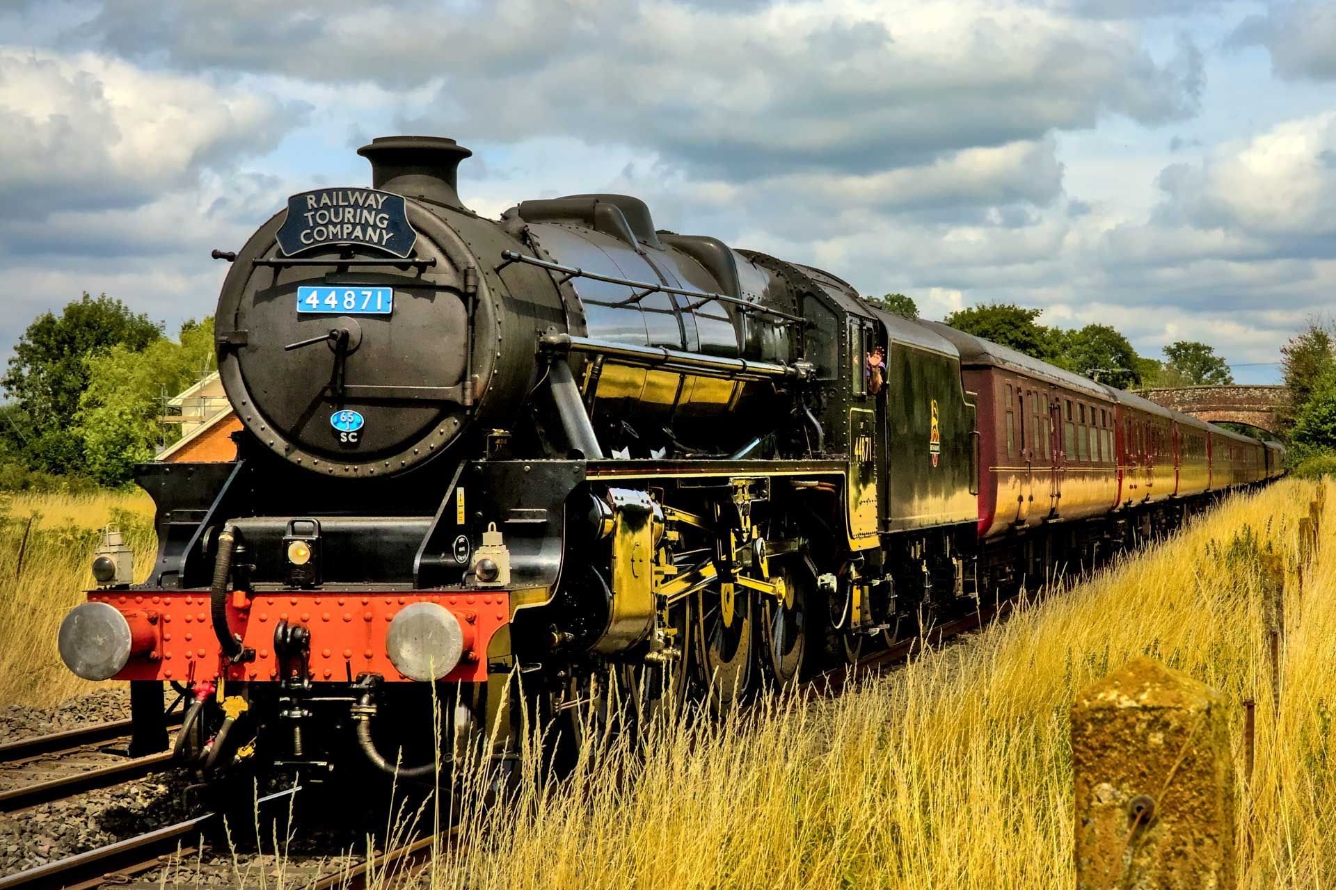 Black steam train with passenger cars on a track, passing through a field with tall grass, under a cloudy sky.