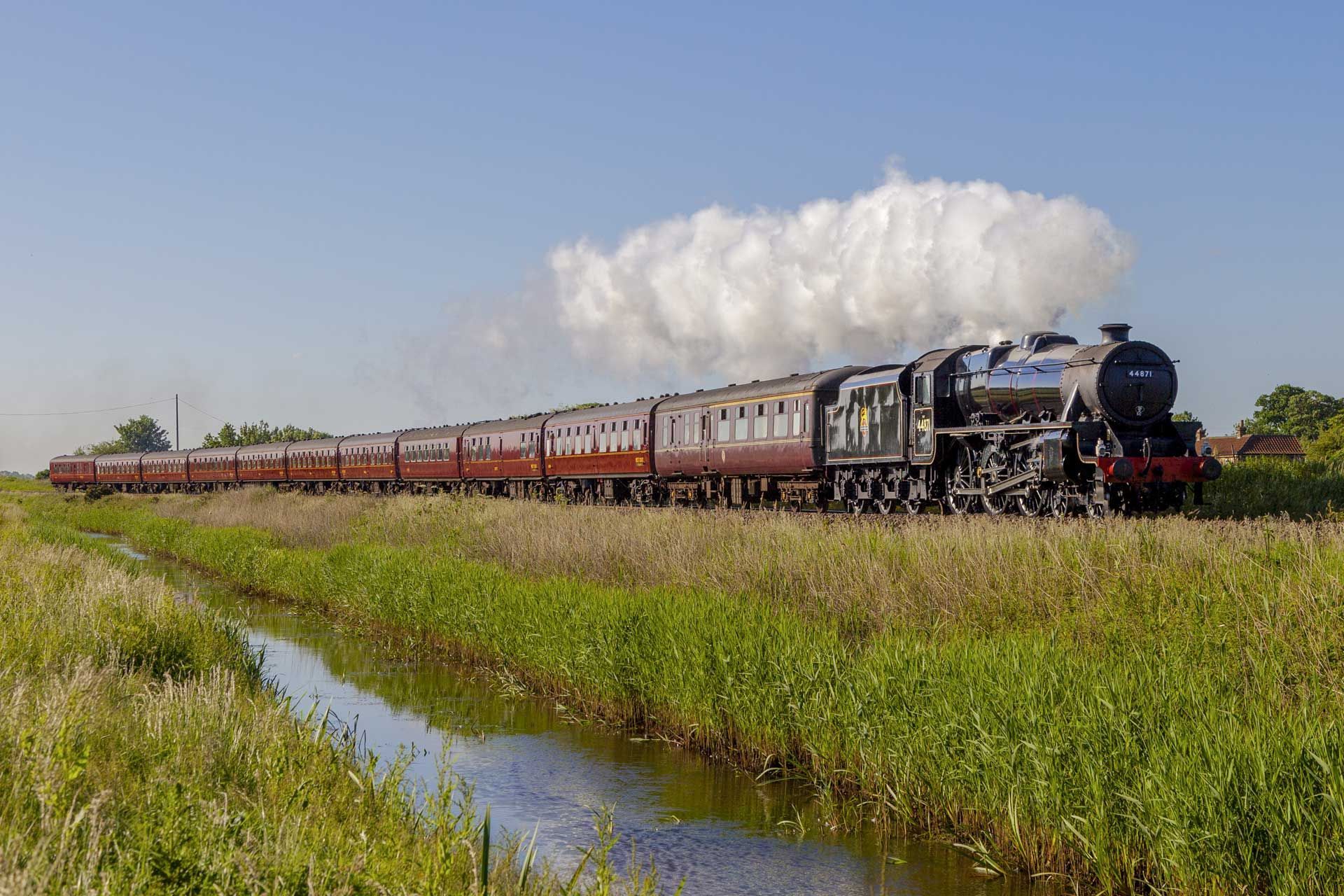 Steam train traveling alongside a canal, emitting smoke against a blue sky.