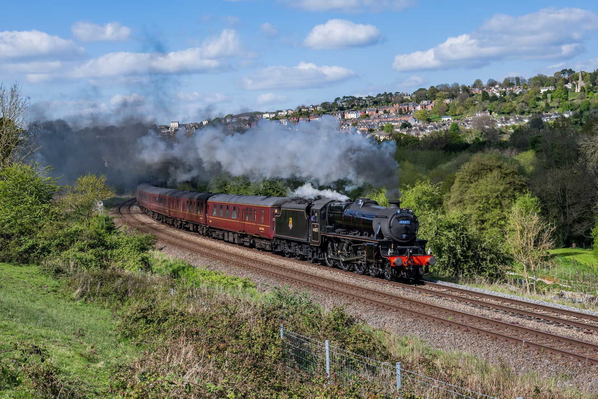 Steam train on tracks curves through a green landscape, smoke billowing. Houses on a hillside in background.