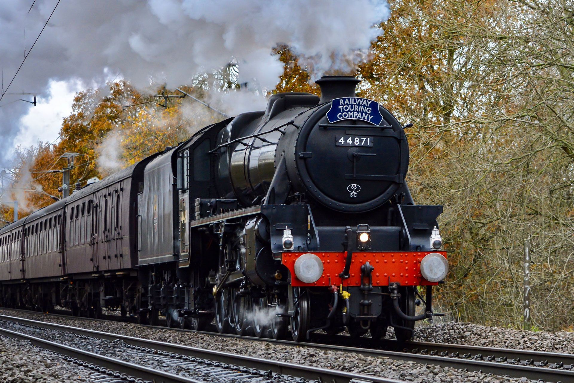 Steam locomotive pulling passenger cars through a wooded autumn landscape
