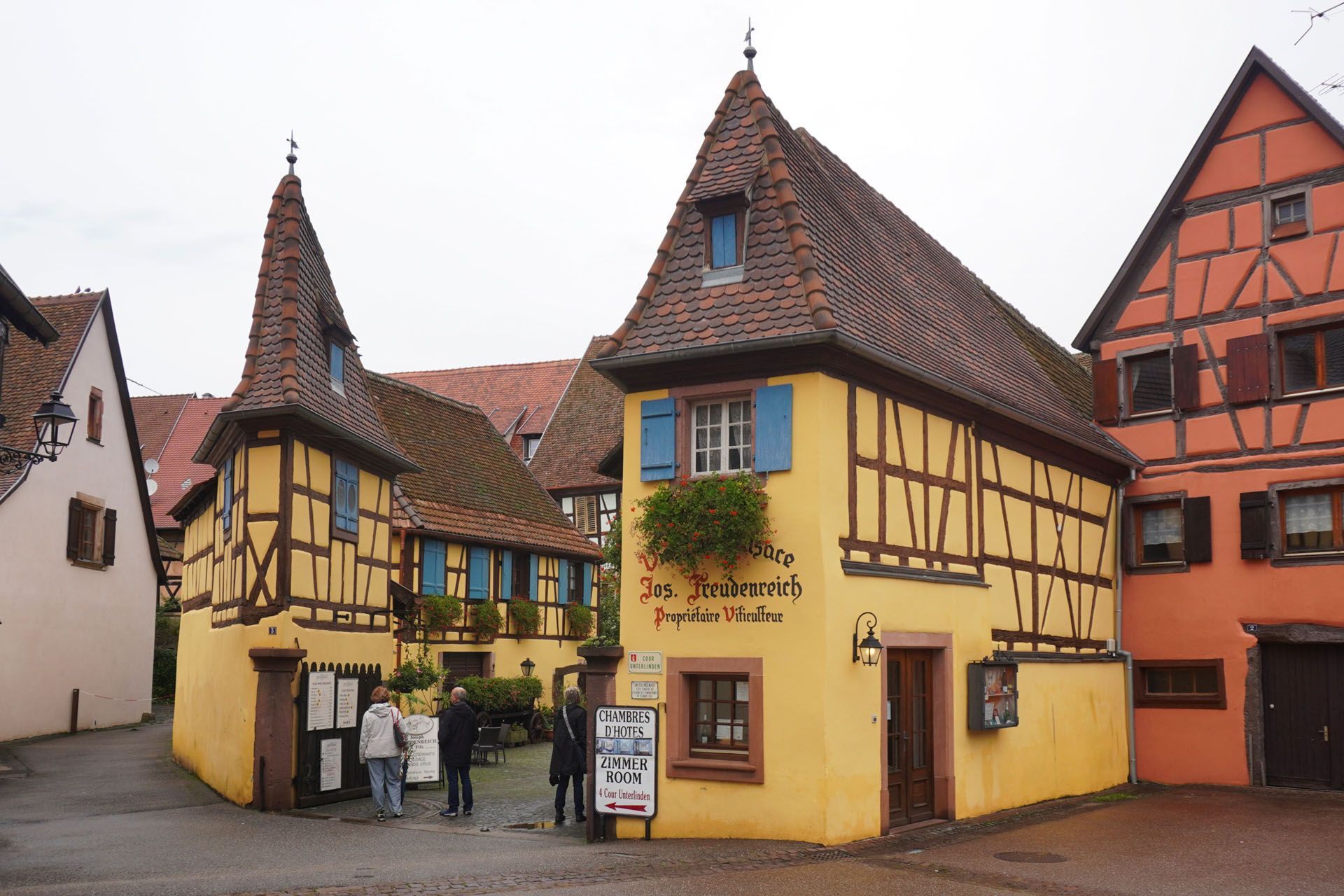 Yellow half-timbered buildings with brown roofs, blue shutters, and pedestrians in a European town square.