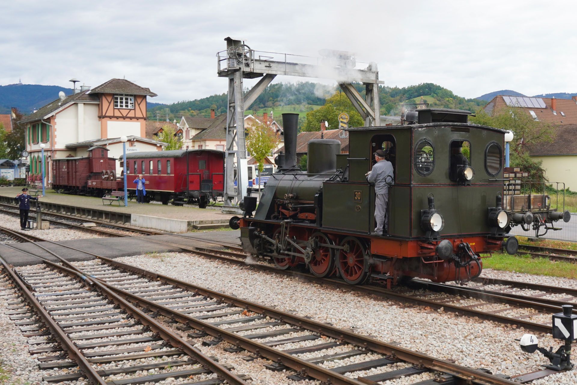 Steam train at a station with a guard, red and brown cars, and a signal.