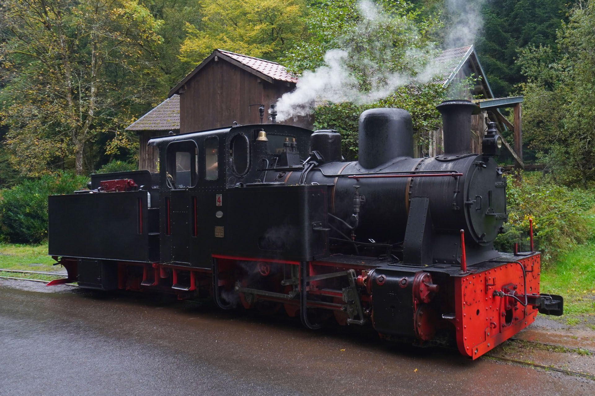 Black and red steam locomotive puffs smoke, parked near a small brown building, set in a wooded area.