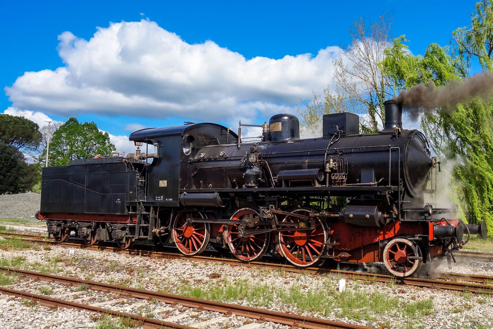 Black steam locomotive on tracks, smoke billowing, red wheels, against a blue sky with clouds.
