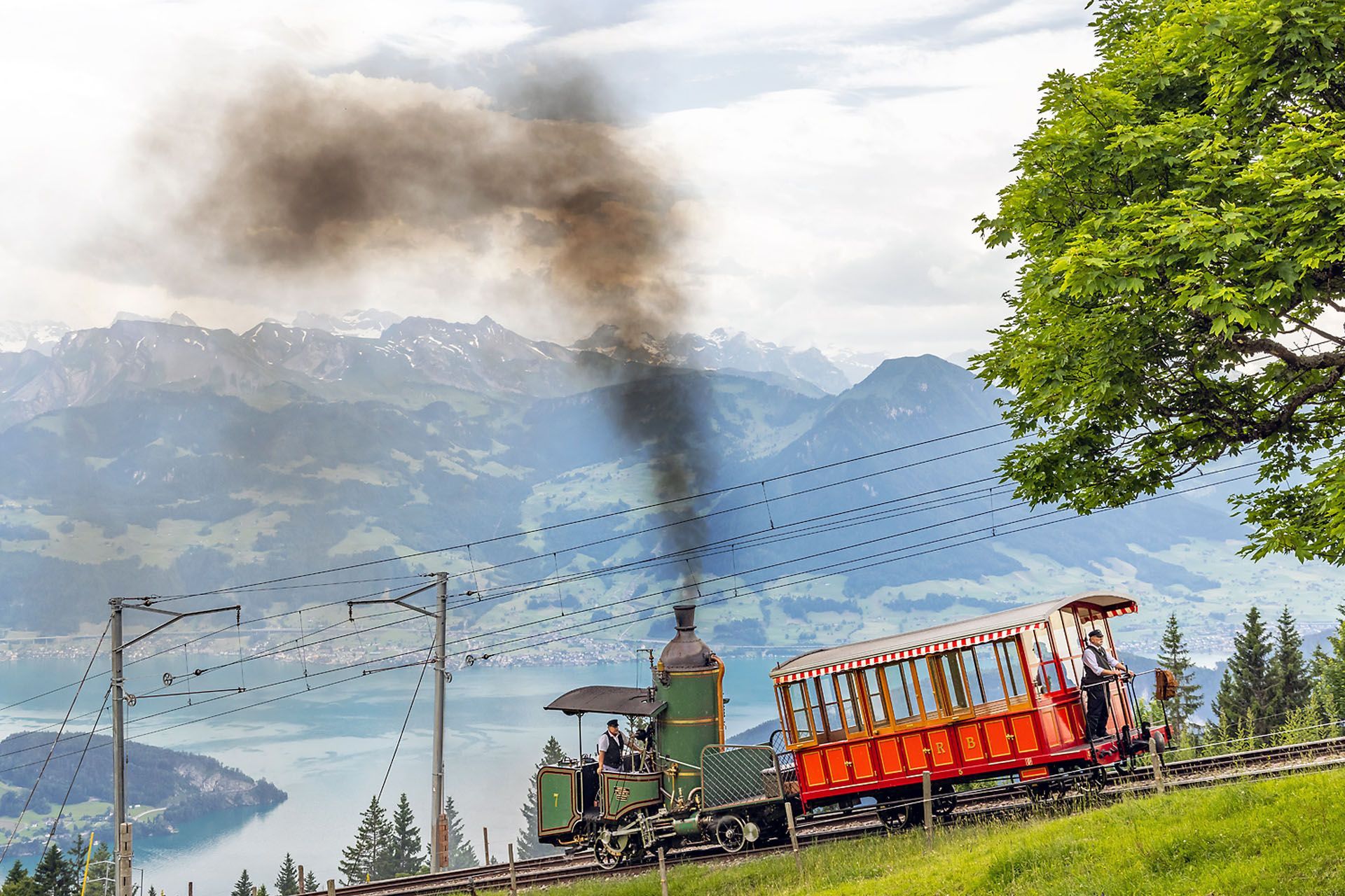 Green steam train ascends a hill, pulling a red passenger car, with a lake and mountains in the background.