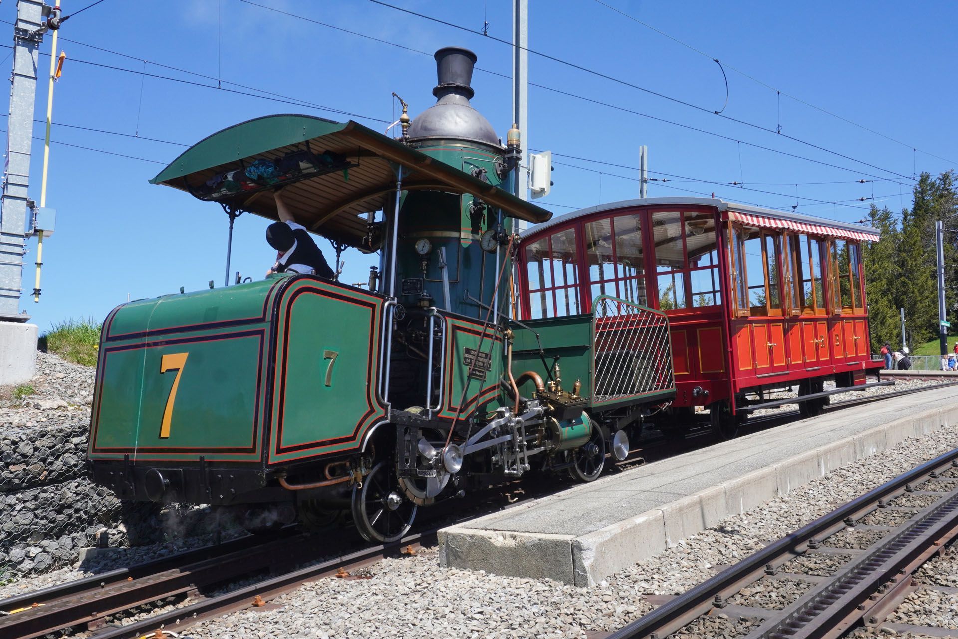 Green and red vintage train, number 7, on tracks, with a driver and open-air car. Sunny day.