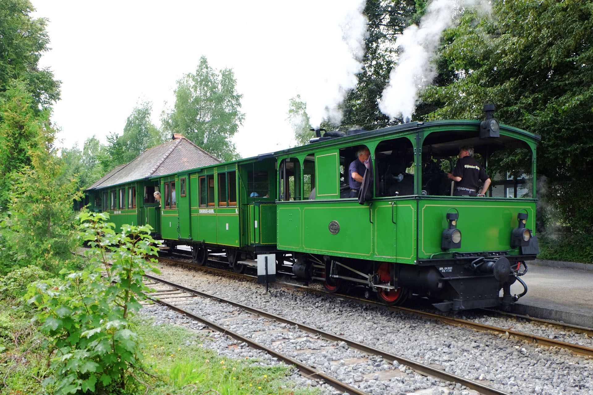 Green steam train on tracks, with passengers and driver. Trees line the sides.