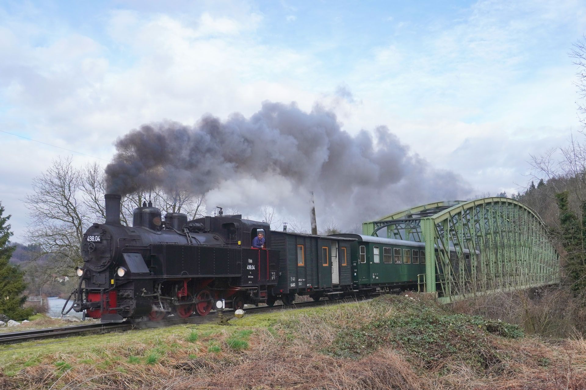 Steam train, black engine with smoke, pulling green passenger cars, passing under a green bridge.