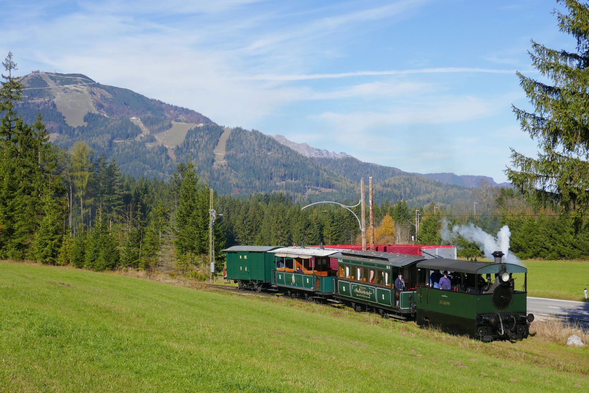 Green train traveling through a grassy field with a mountain backdrop under a blue sky.