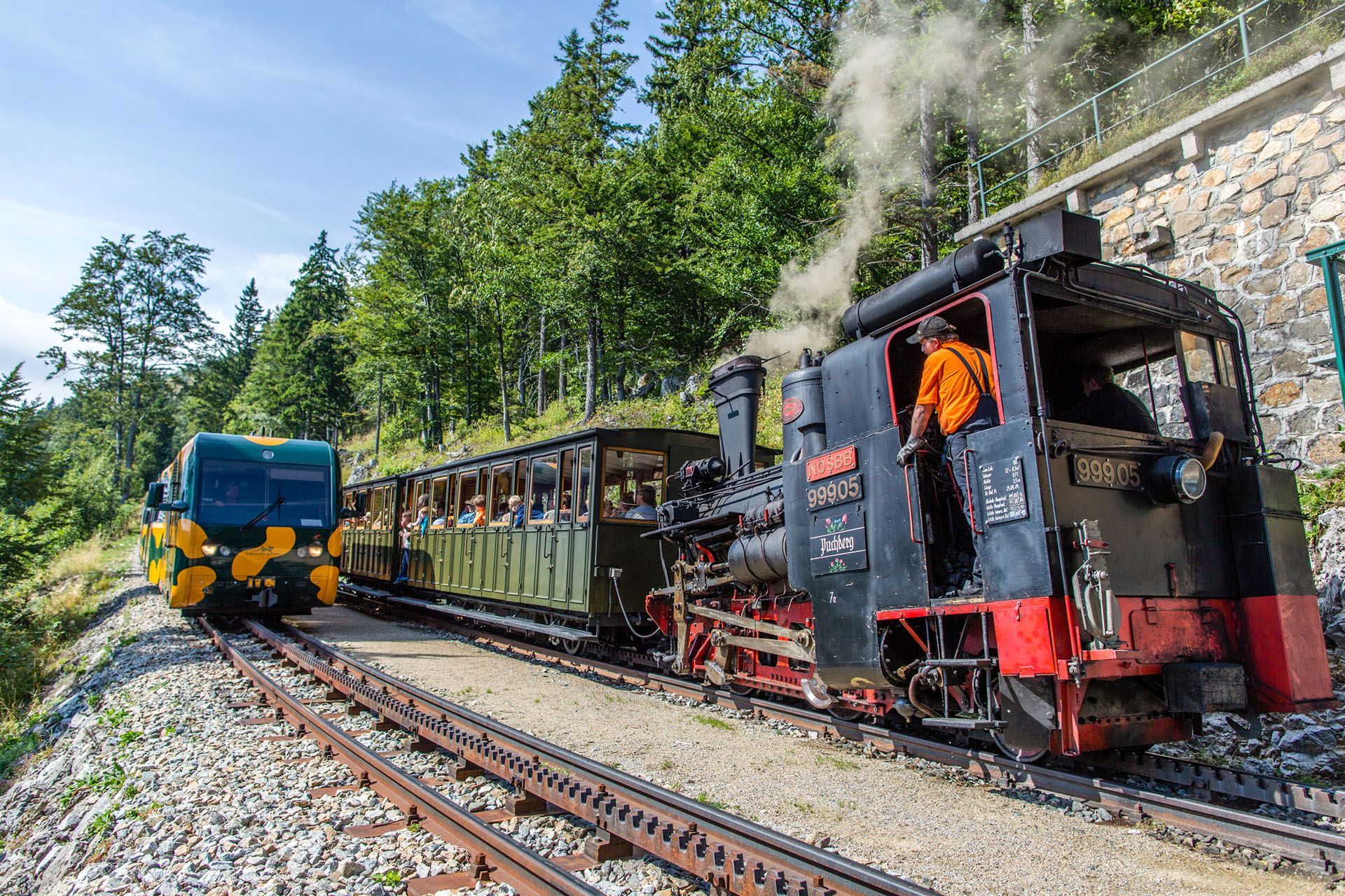Steam train on tracks near a forest; conductor in orange shirt; a green bus-like vehicle.