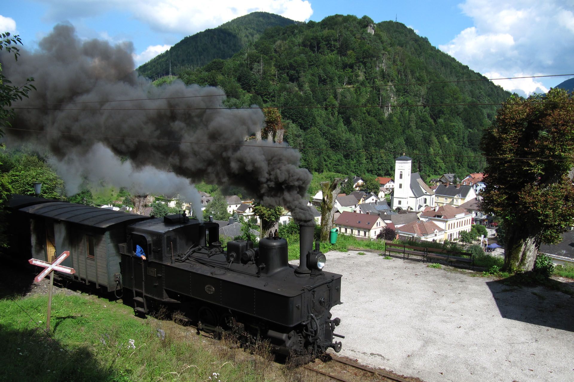 Steam train puffs smoke near a village with a church and green mountain backdrop.