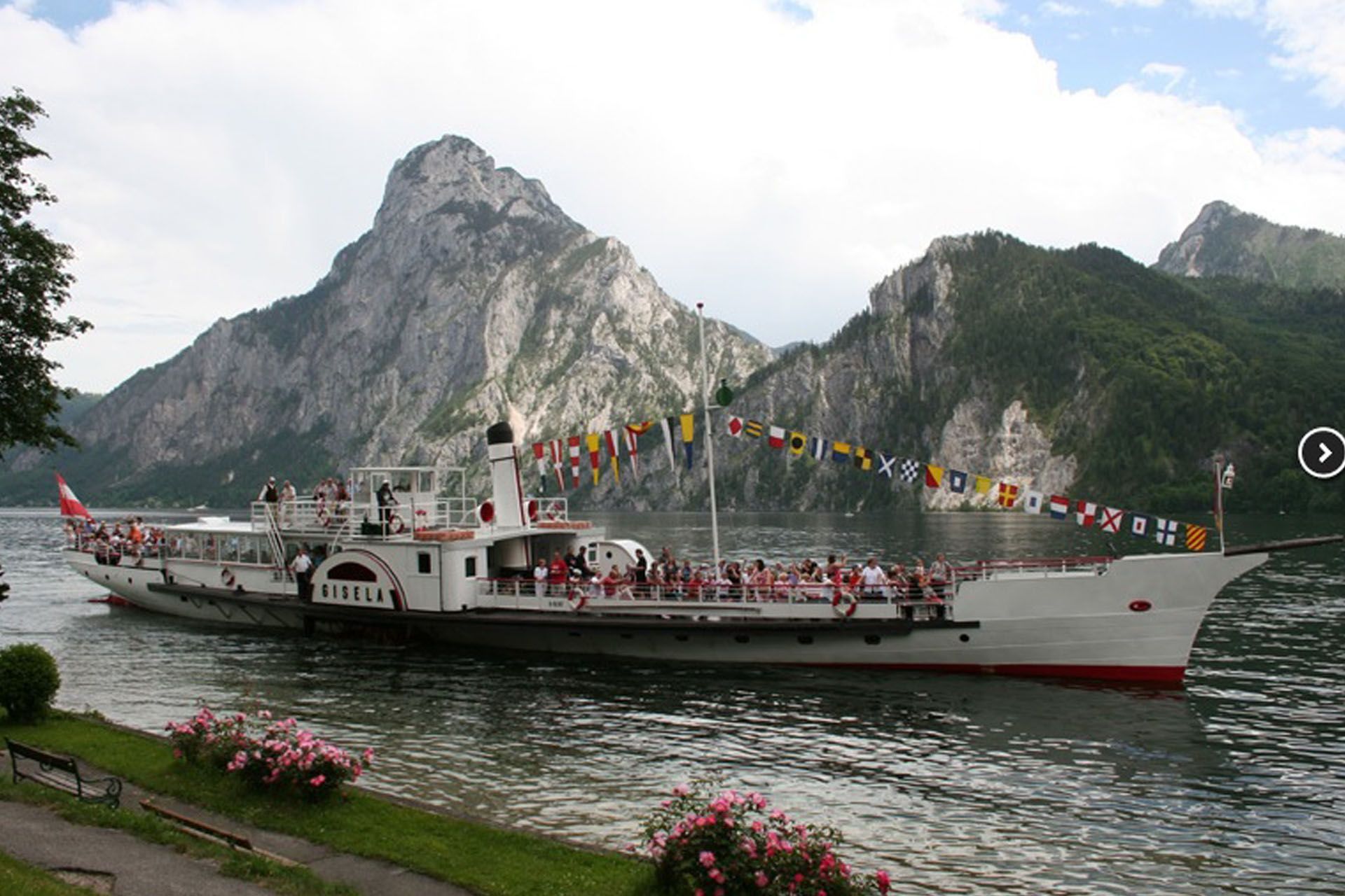 Paddle steamer on a lake, decorated with flags. Mountains in the background. People on deck.
