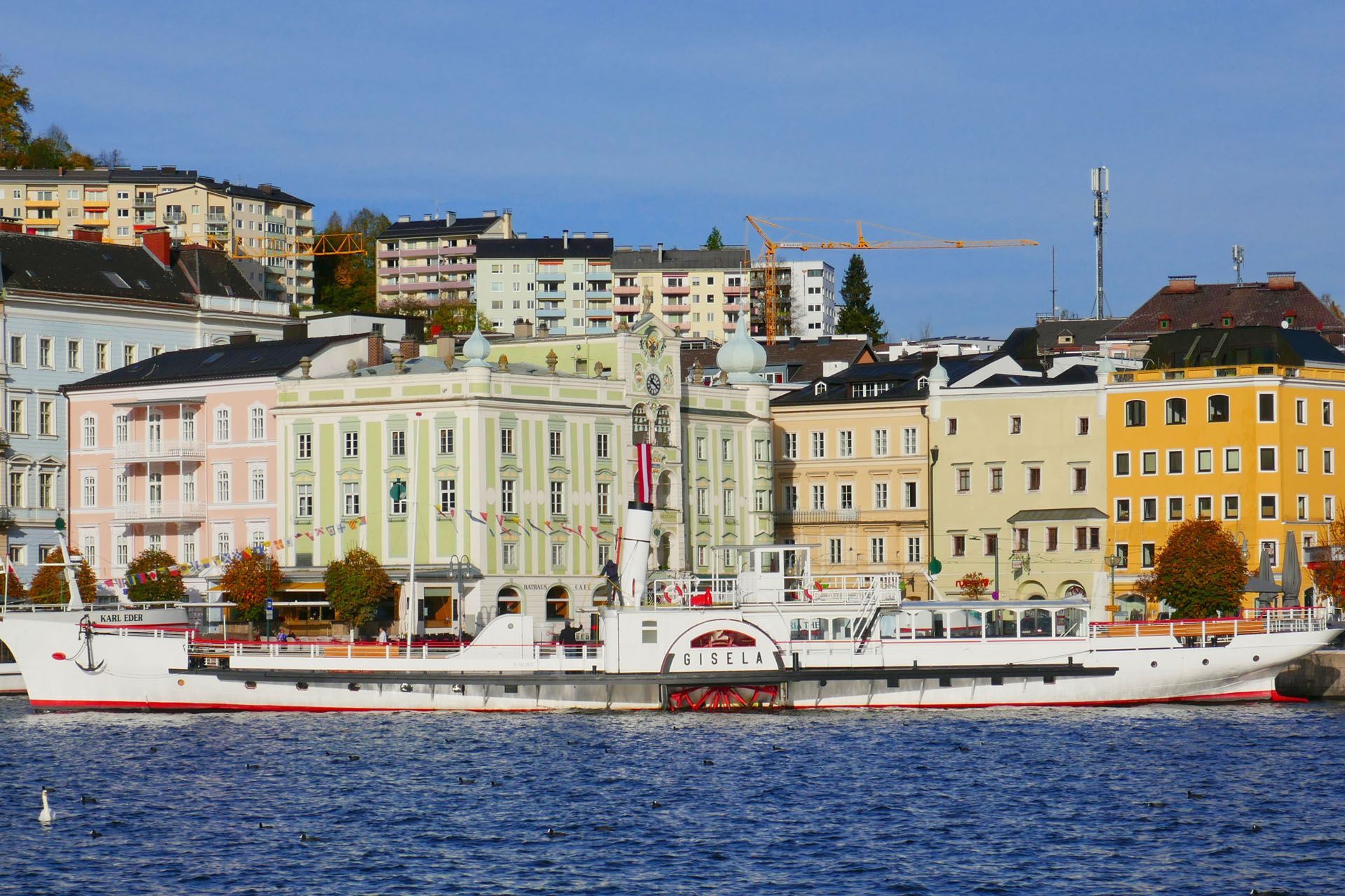 Paddle steamer docked in front of colorful buildings on a waterfront under a blue sky.