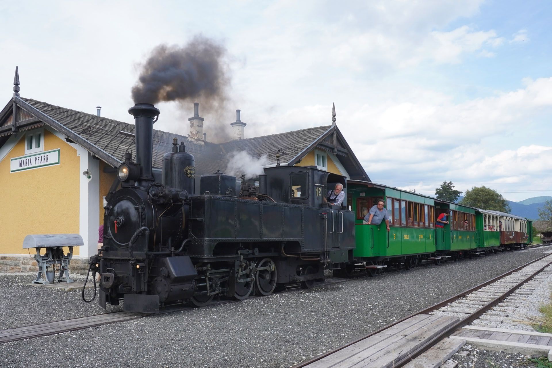 Black steam train pulling green and red passenger cars at a yellow station with people inside.
