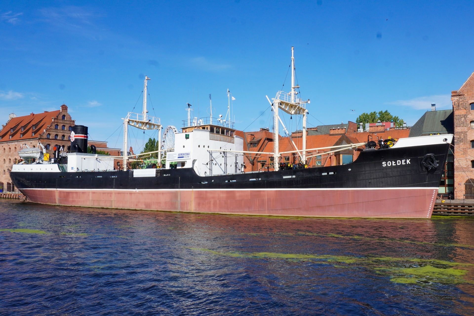 Large black and red ship docked in a canal, with buildings in the background under a blue sky.