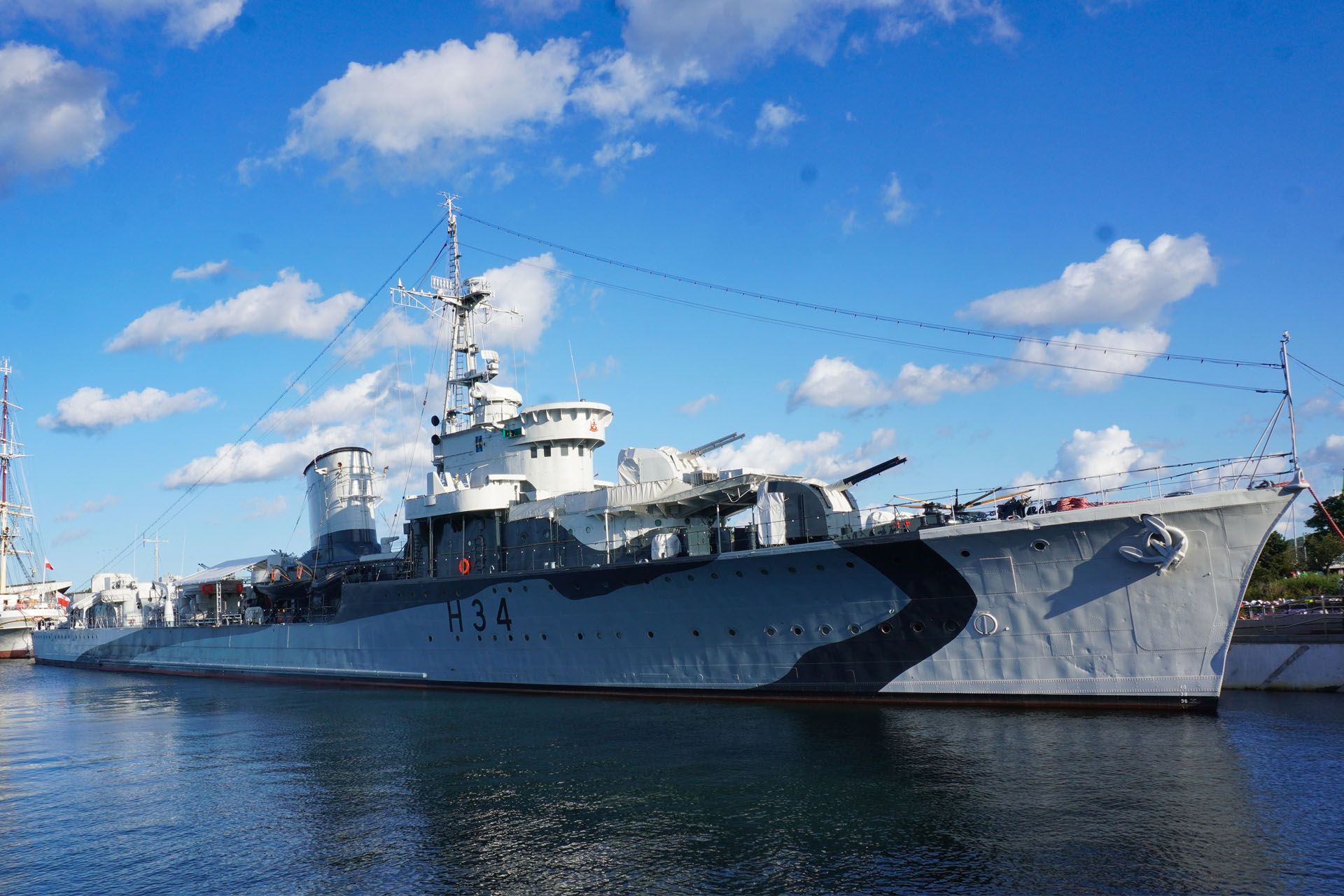 Warship, gray and black camouflage, docked in water, under a blue sky with clouds.