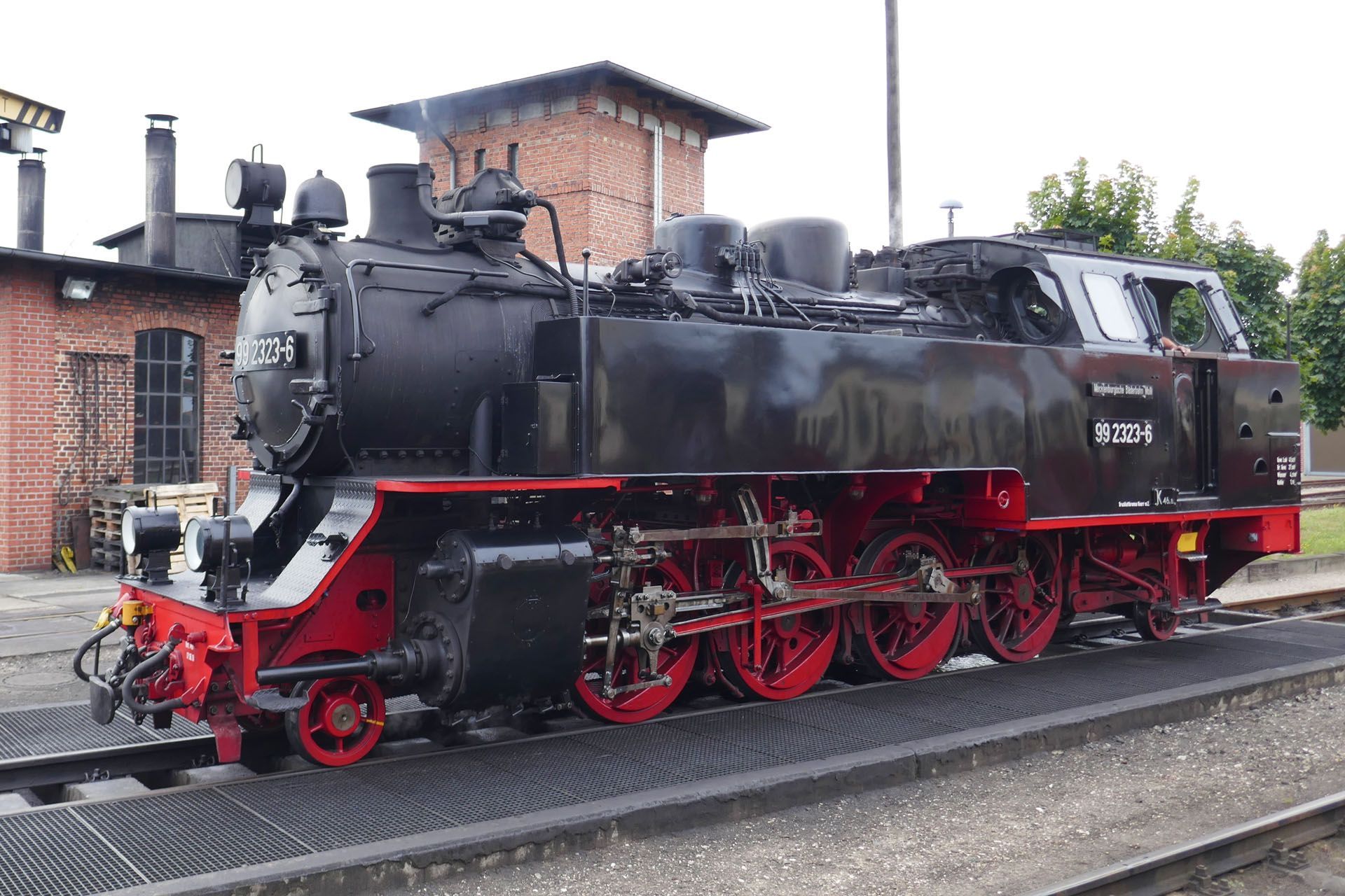 Black steam locomotive on tracks, red wheels, building in background.
