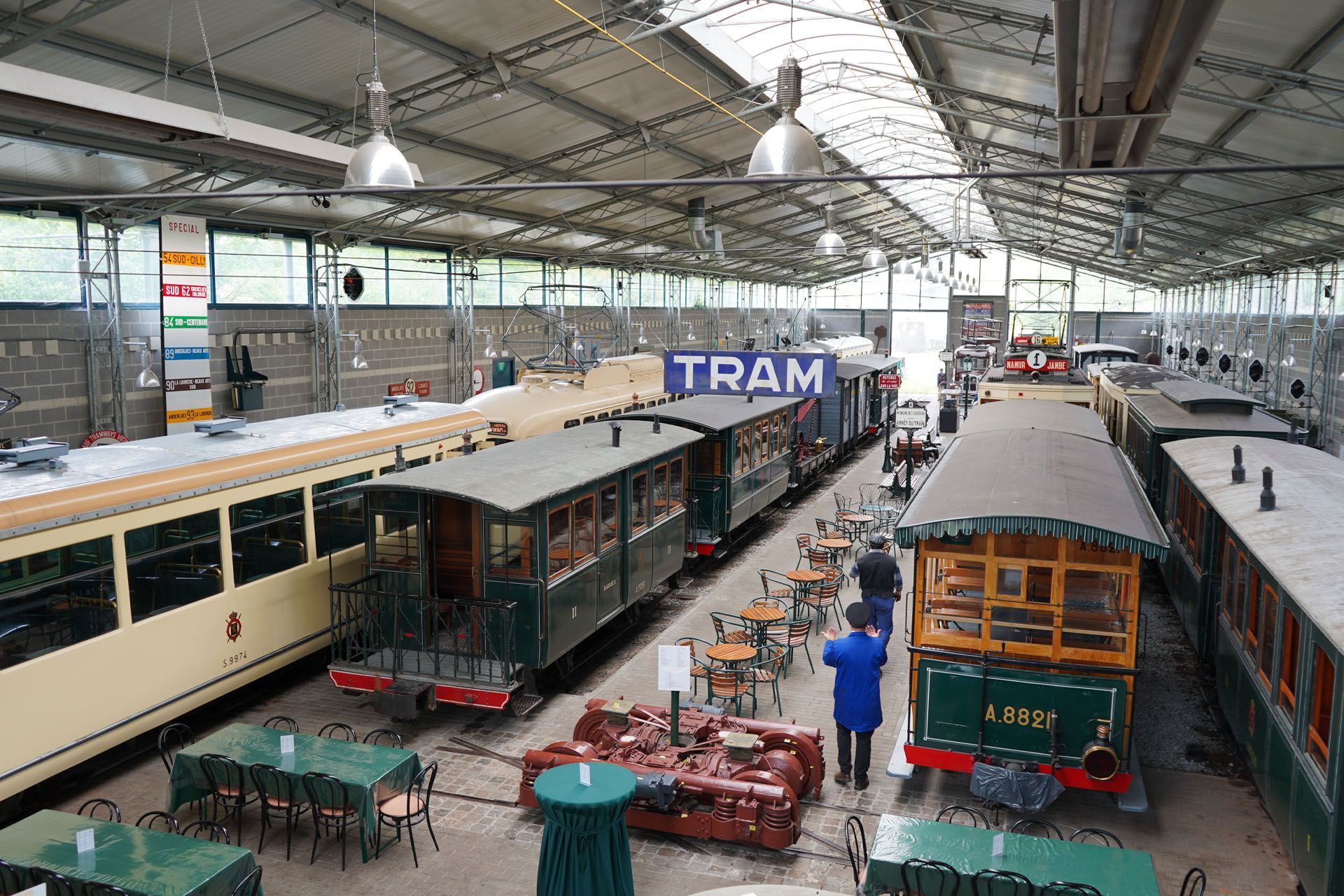 Inside a tram museum, several vintage trams are displayed. People walk around and tables are set up.