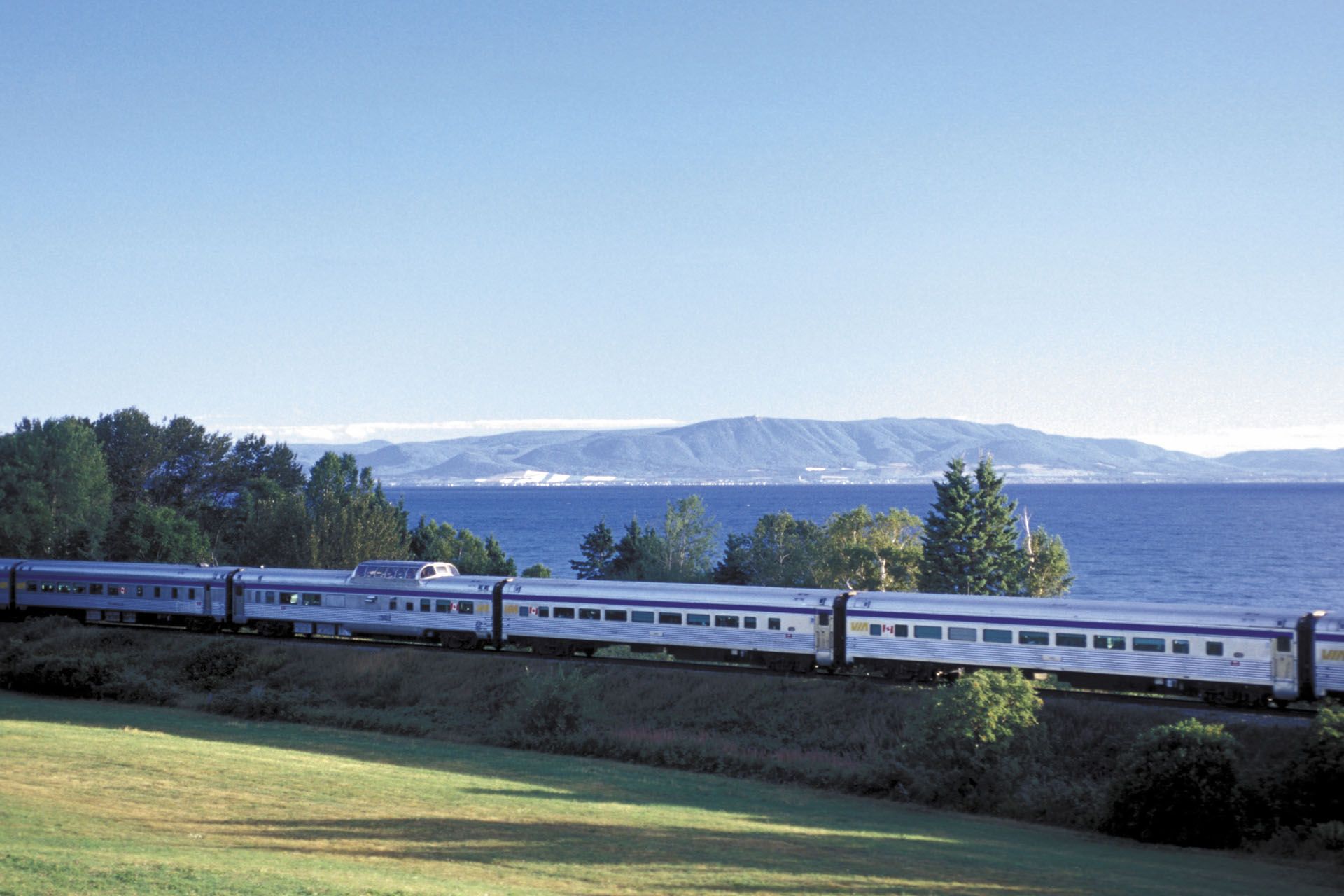 Train traveling alongside a body of water with a mountain range in the distance on a sunny day.