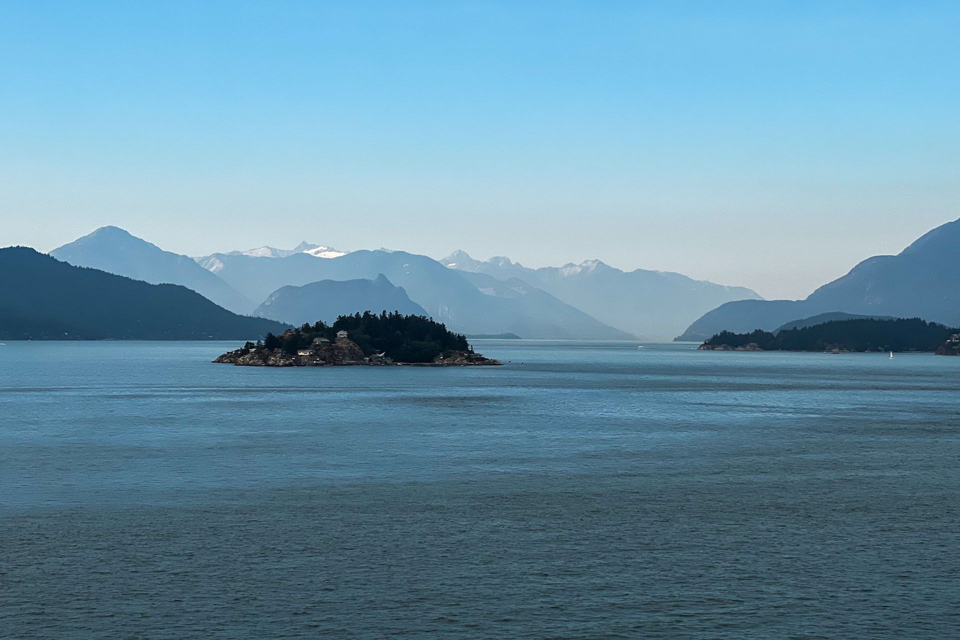 Island in a bay with mountains in the background, under a blue sky; water in the foreground.