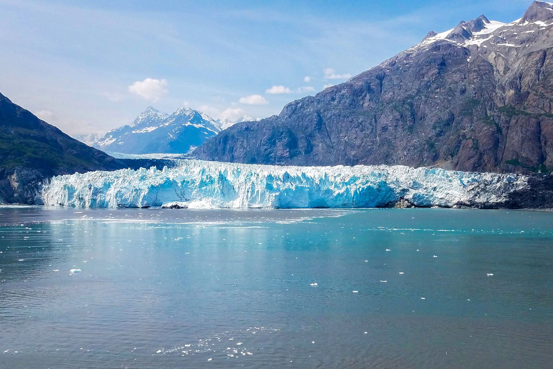 Glacier meets water, backed by mountains. Bright blue ice reflects in the sea under a blue sky.