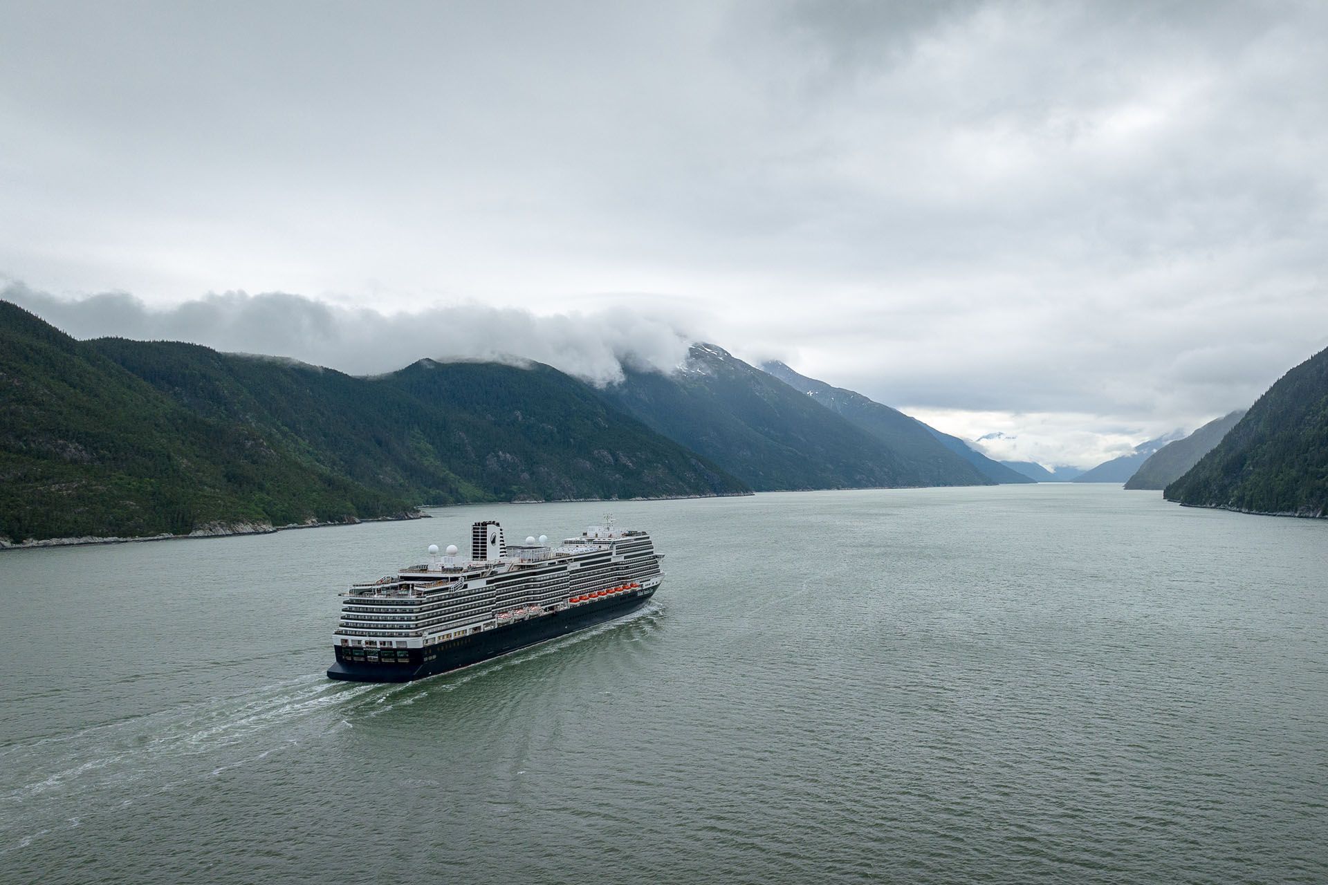 Large cargo ship sailing through a fjord, surrounded by mountains under a cloudy sky.